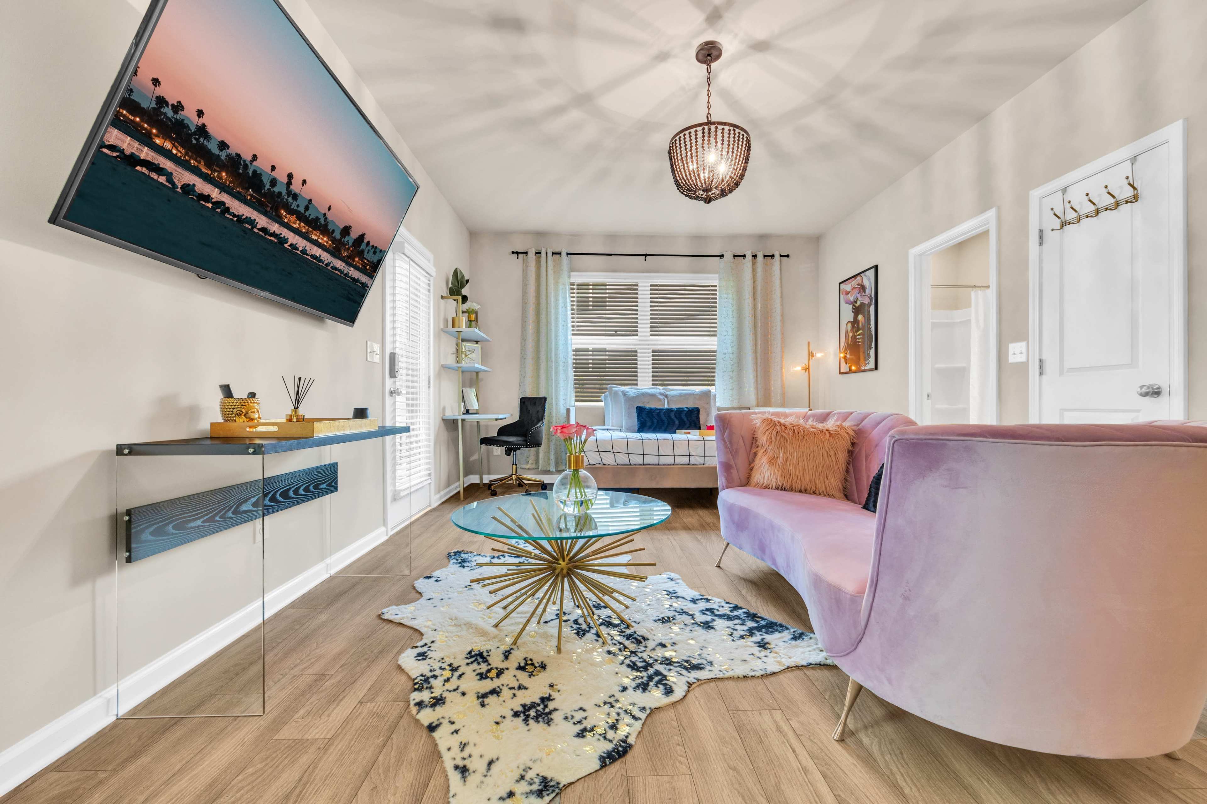 The image shows a modern living room featuring a pink velvet sofa, a glass coffee table on a patterned rug, a wall-mounted TV, and a window with curtains.