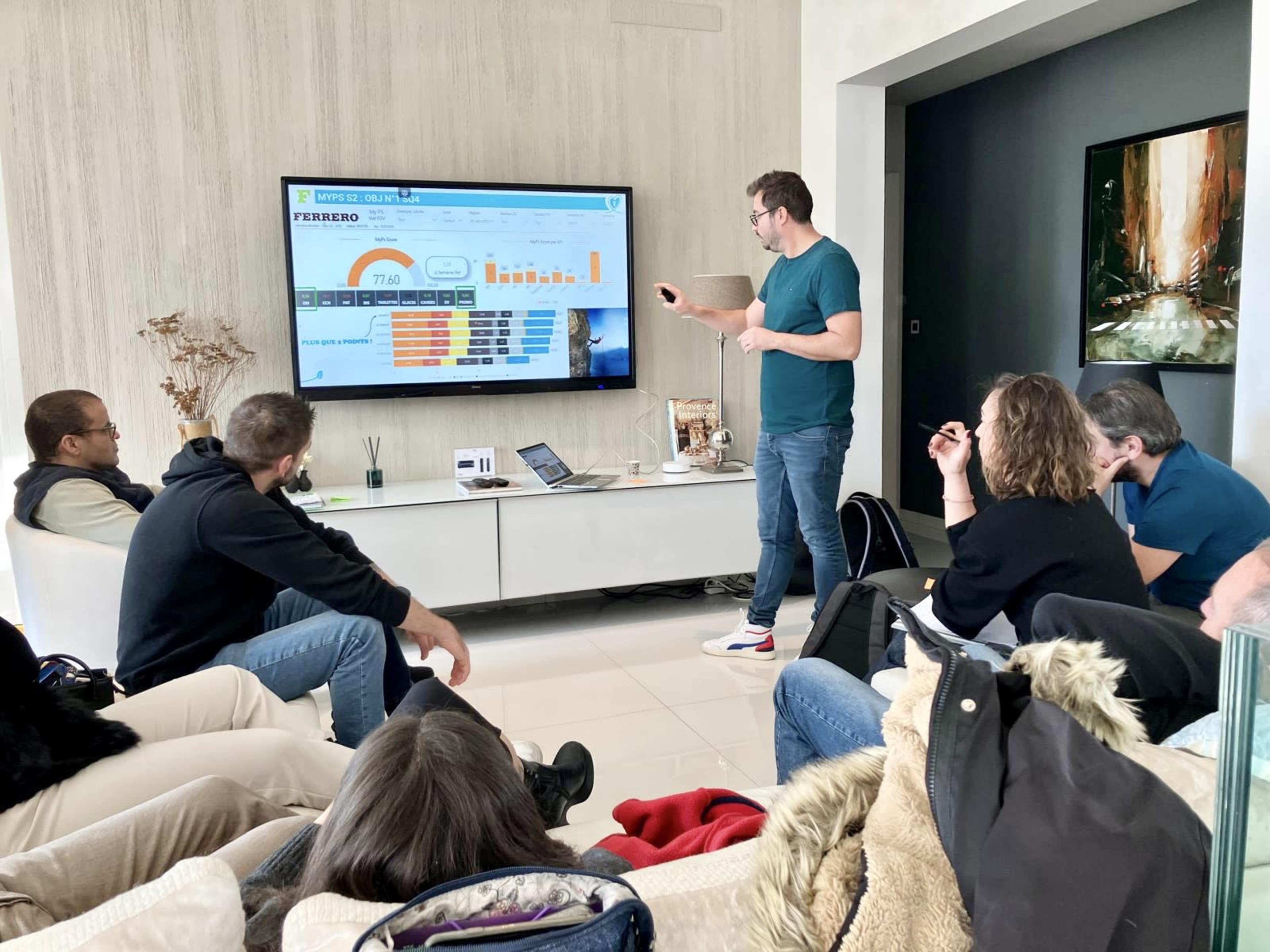 A man stands in front of a large screen, presenting data to a group of seated individuals in a modern living room.