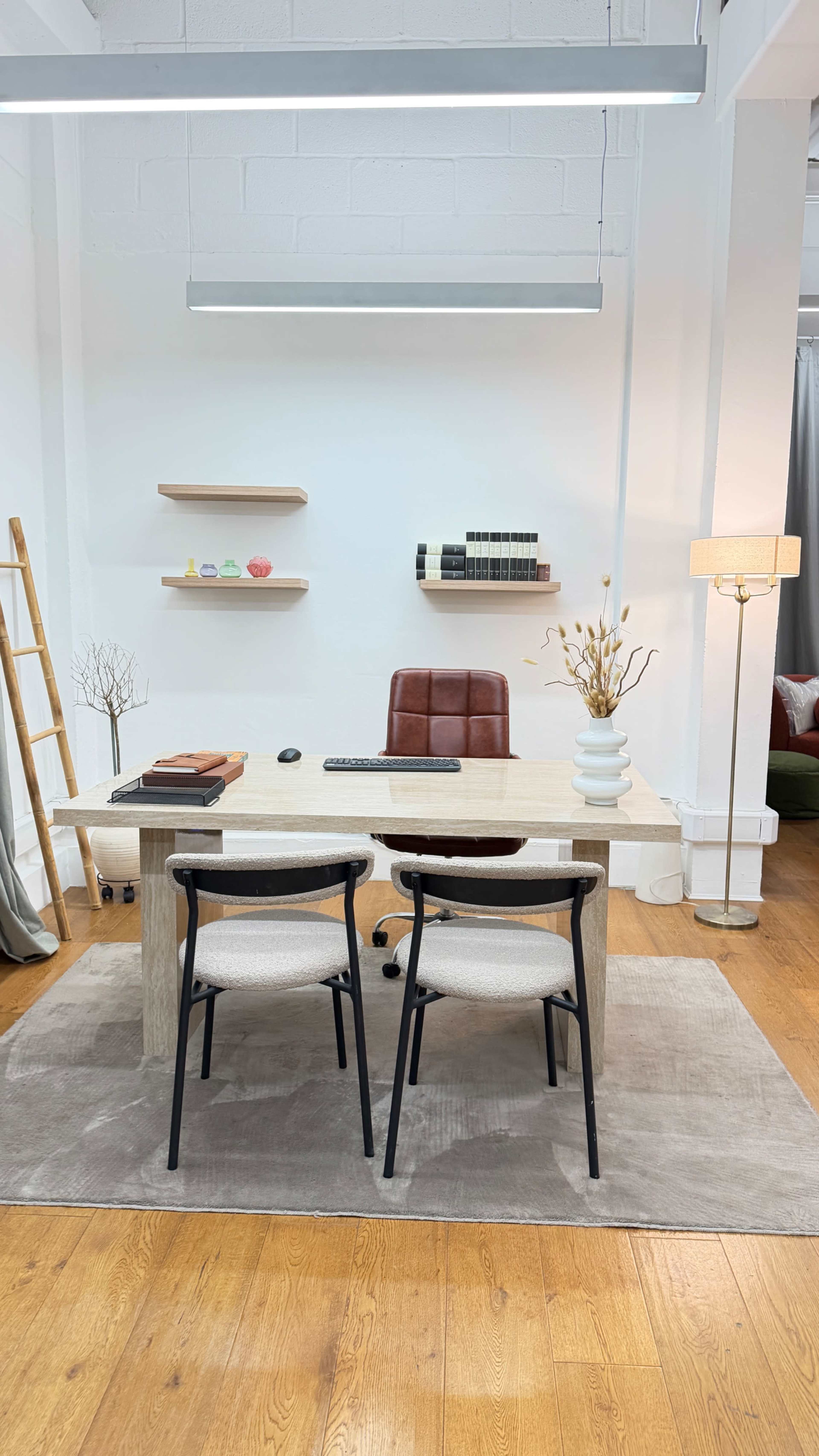 A modern office space features a wooden desk with a computer and two chairs, set against a simple white wall with decorative shelves.