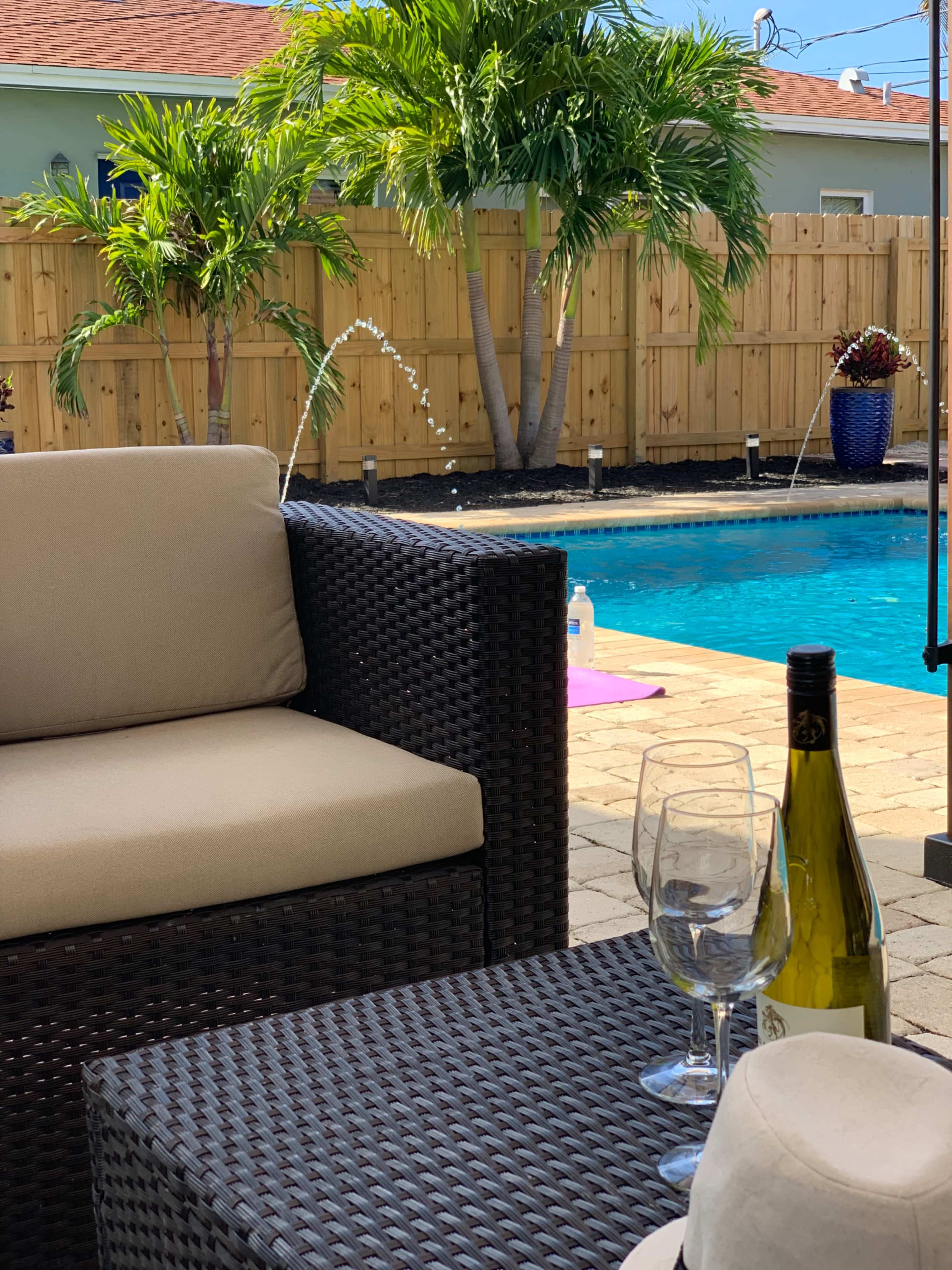 A patio seating area with a wicker couch and table overlooks a swimming pool surrounded by landscaping and palm trees.