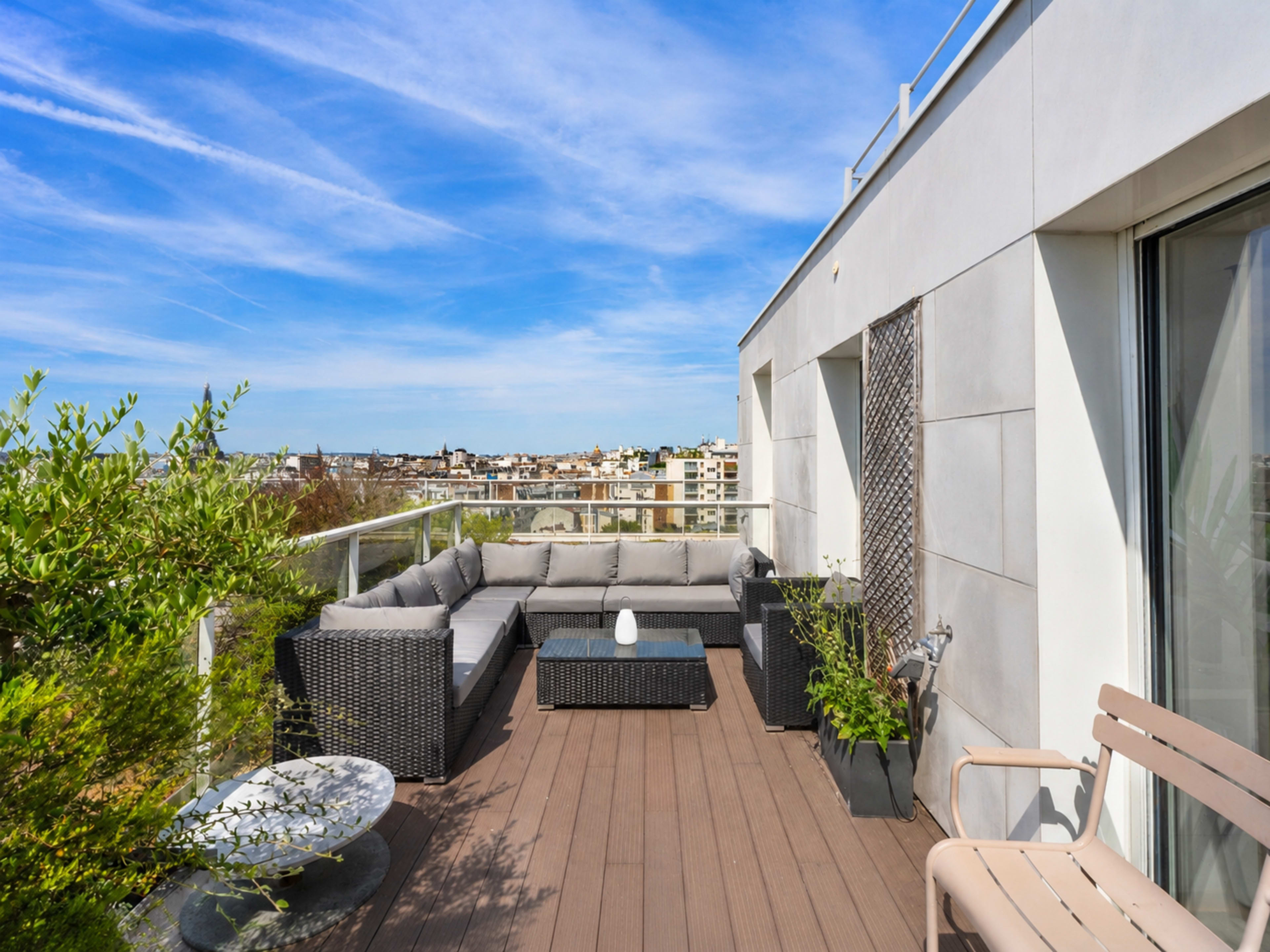 The image shows a modern terrace with a seating area comprised of gray sofas and a coffee table, overlooking a city skyline.