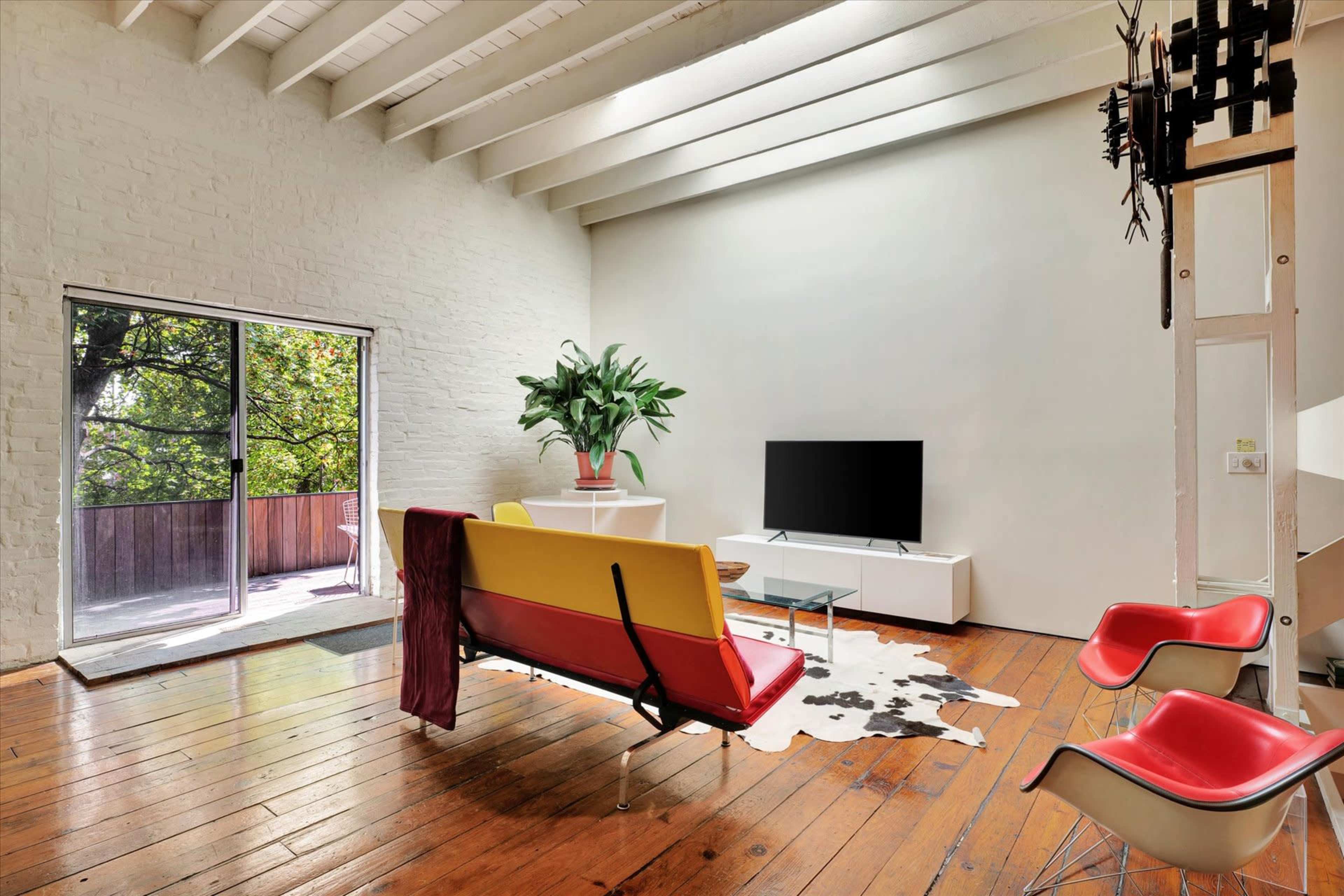 The image shows a modern living room with a yellow and red sofa, wooden floors, a potted plant, a television on a white cabinet, and a sliding glass door leading to a balcony.