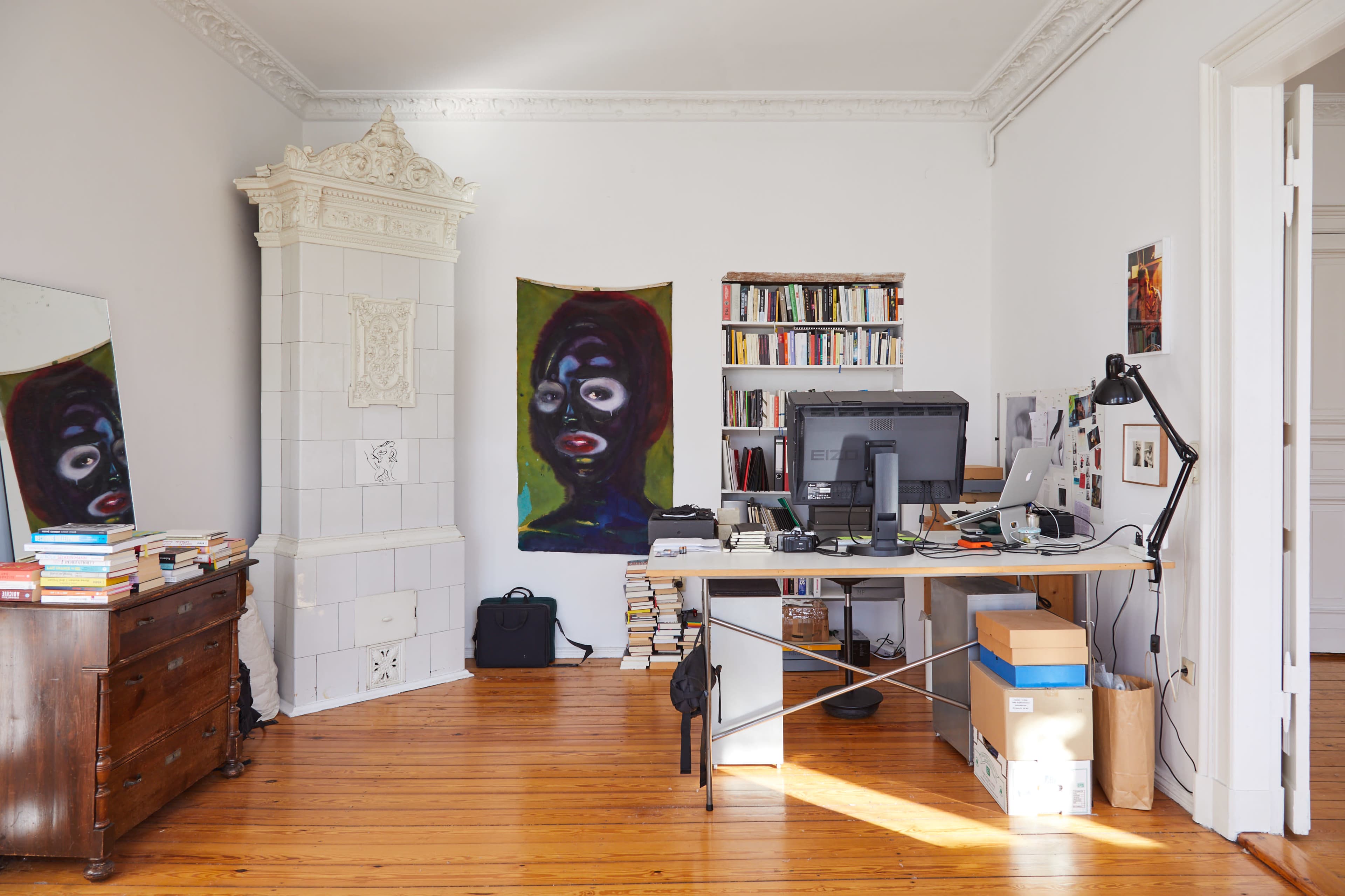 The image shows a tidy office space featuring a desk with a computer, a bookshelf filled with books, a large painting on the wall, and a decorative fireplace.