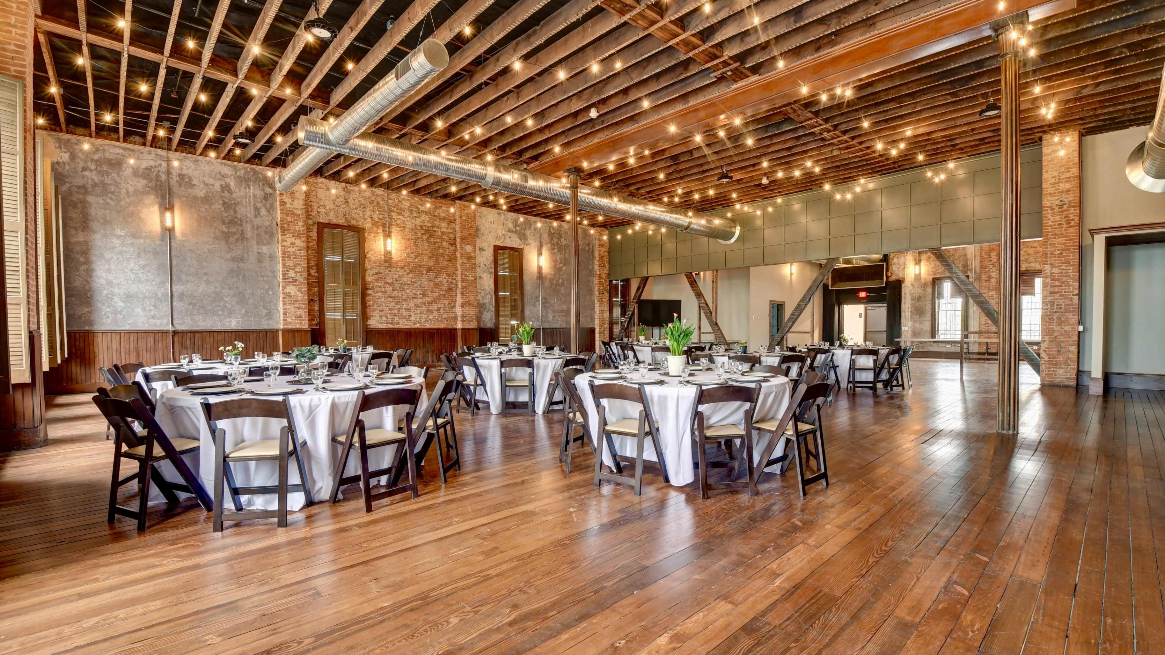 A spacious dining area featuring several round tables set with white tablecloths and decorative centerpieces, under a ceiling with exposed beams and string lights.