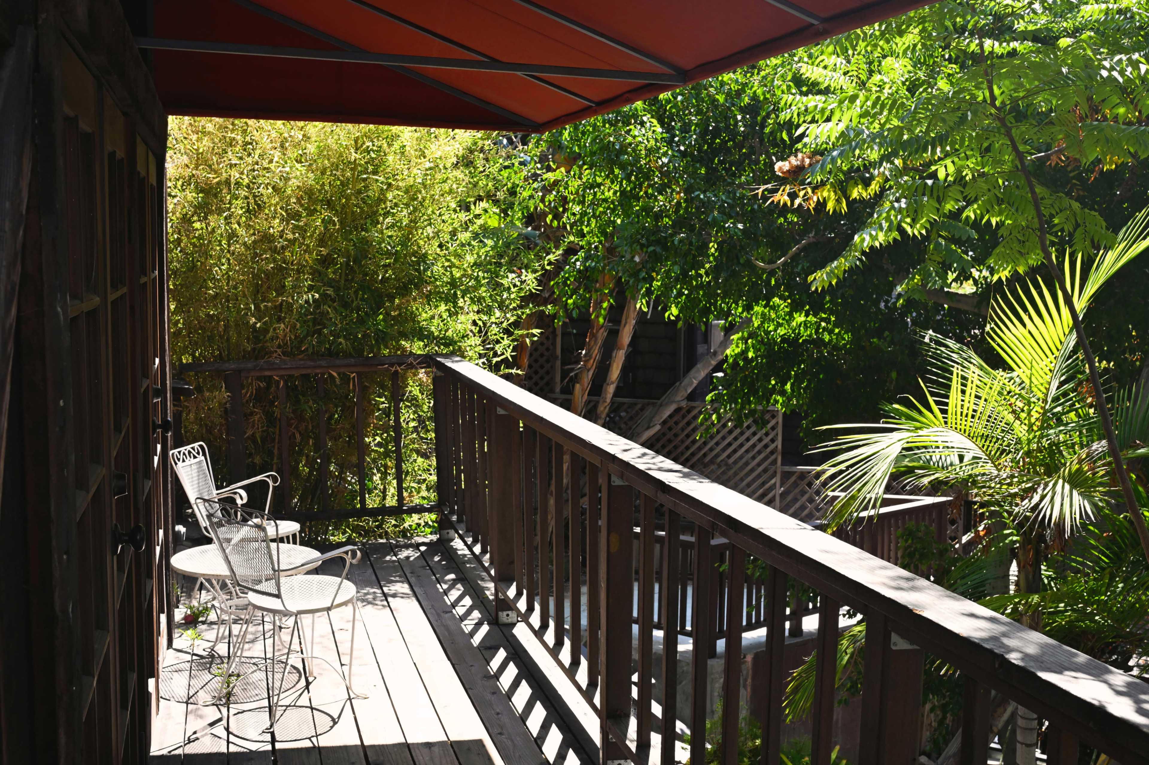 A wooden balcony features two metal chairs overlooking a lush, green garden filled with various plants and trees.