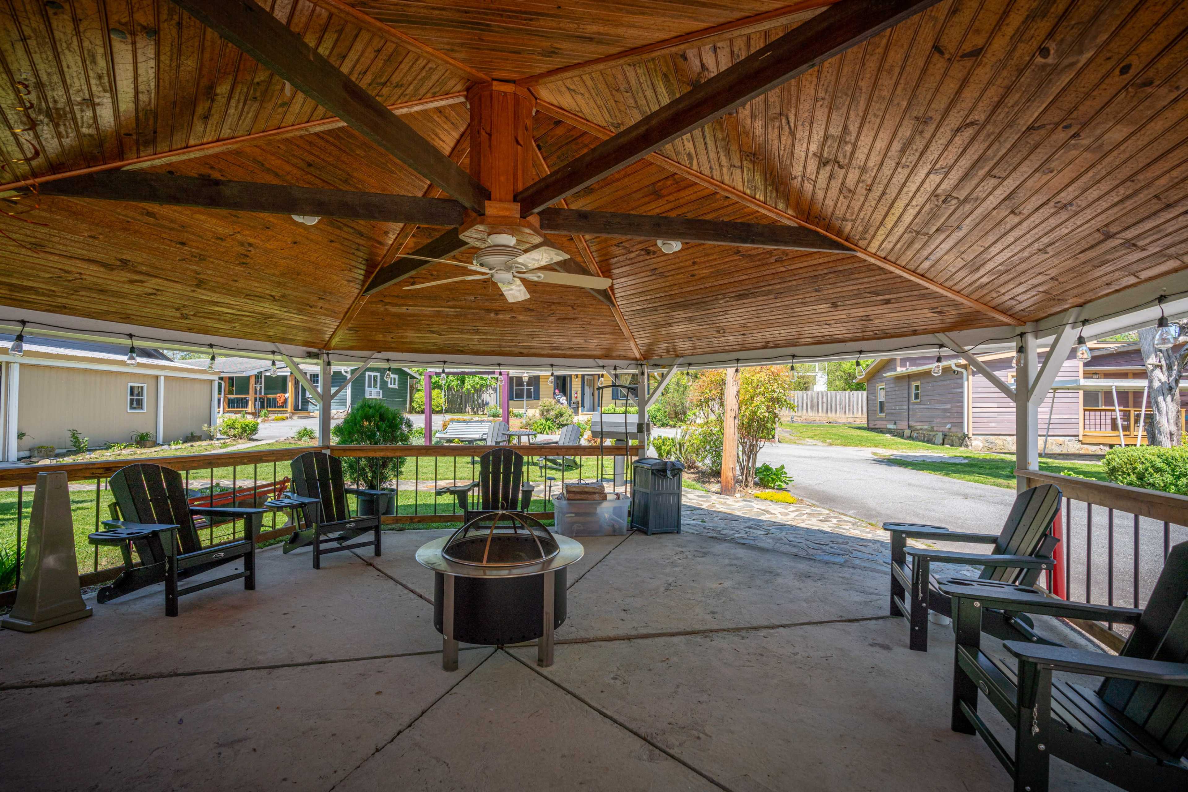 A wooden gazebo with a circular seating arrangement, a fire pit in the center, and a view of a landscaped area beyond.