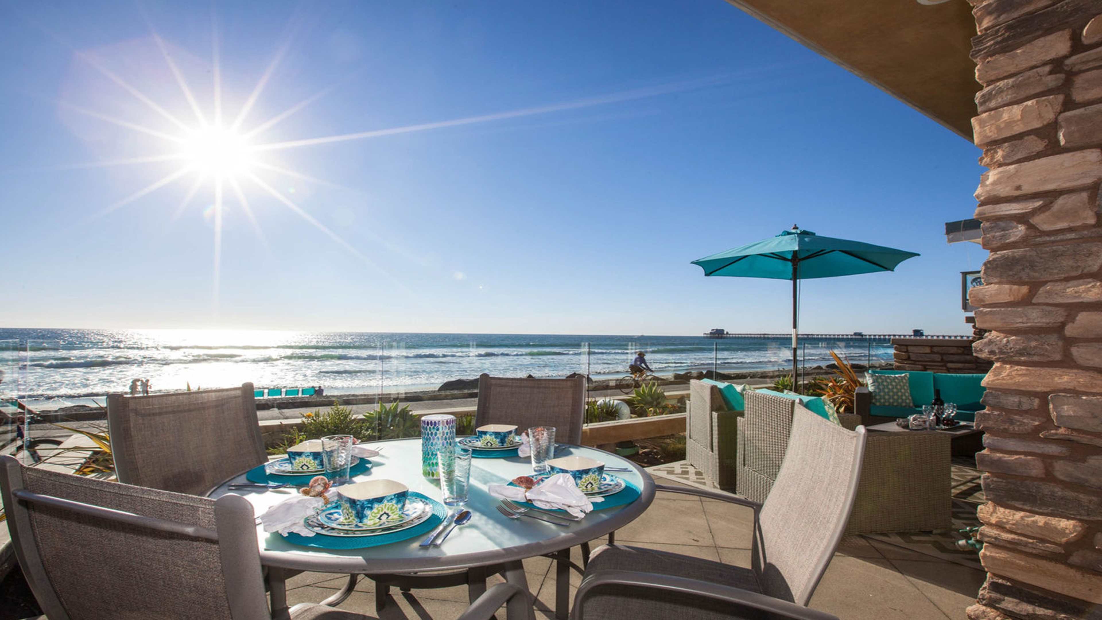 A dining table is set on a patio overlooking the ocean, with waves crashing in the background and a bright sun shining in the sky.