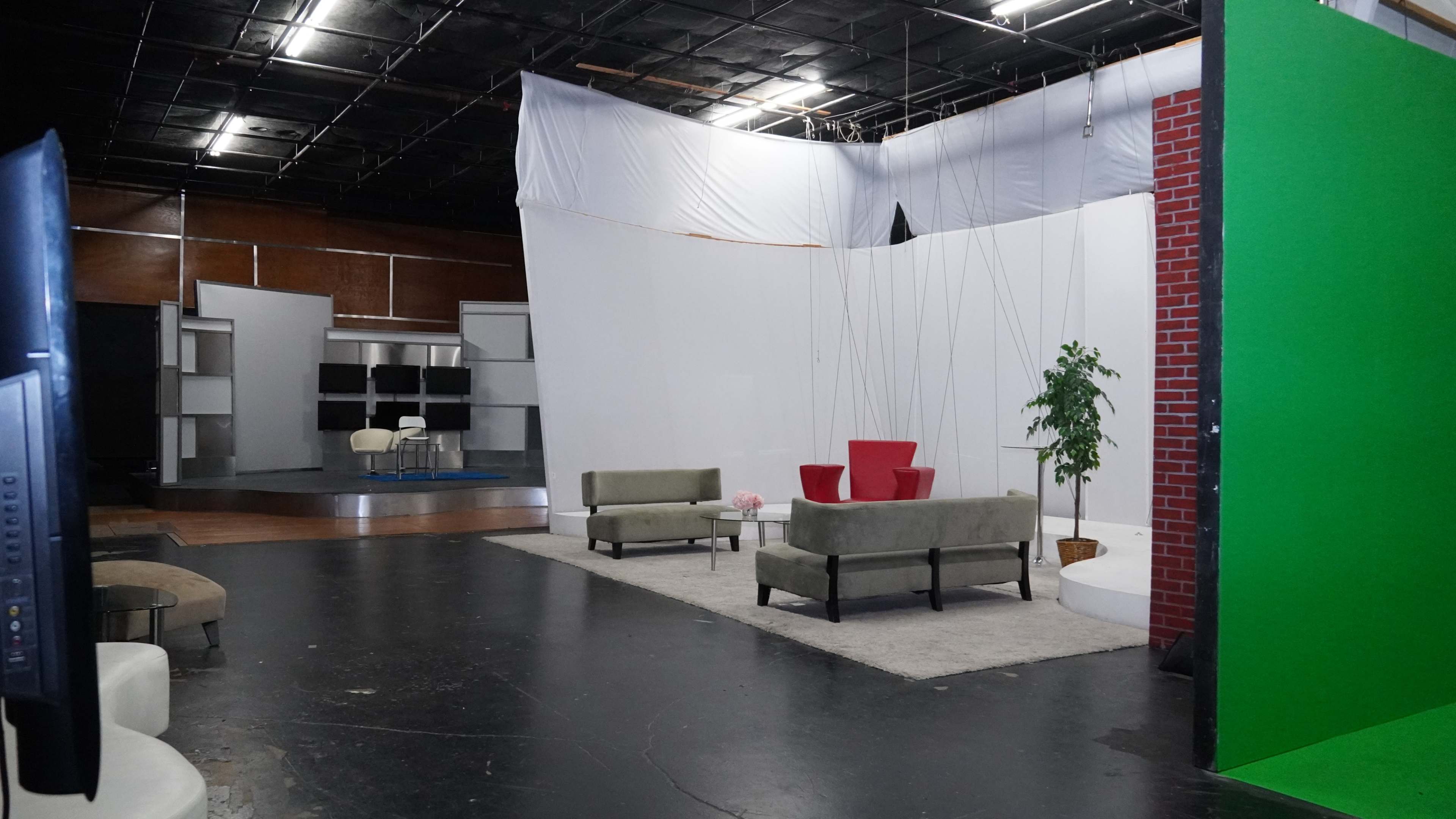 The image shows a television studio with a set featuring a couple of green sofas, a red chair, a small table, and a backdrop with a green screen in one corner.