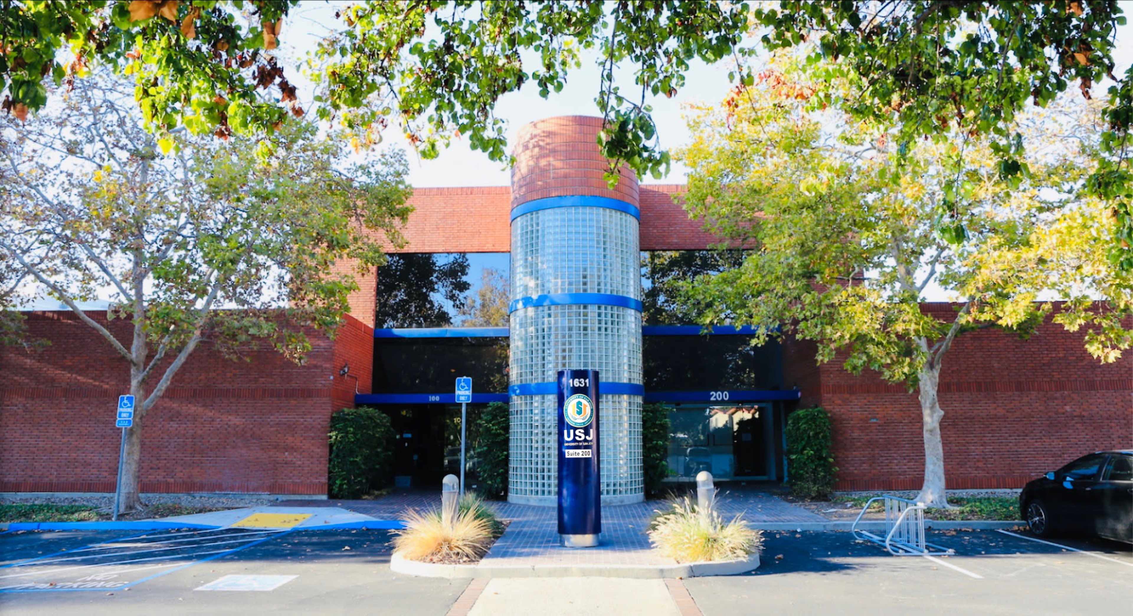 The image shows a modern brick building with a glass entrance, flanked by trees and a blue sign indicating the U.S. Postal Service.