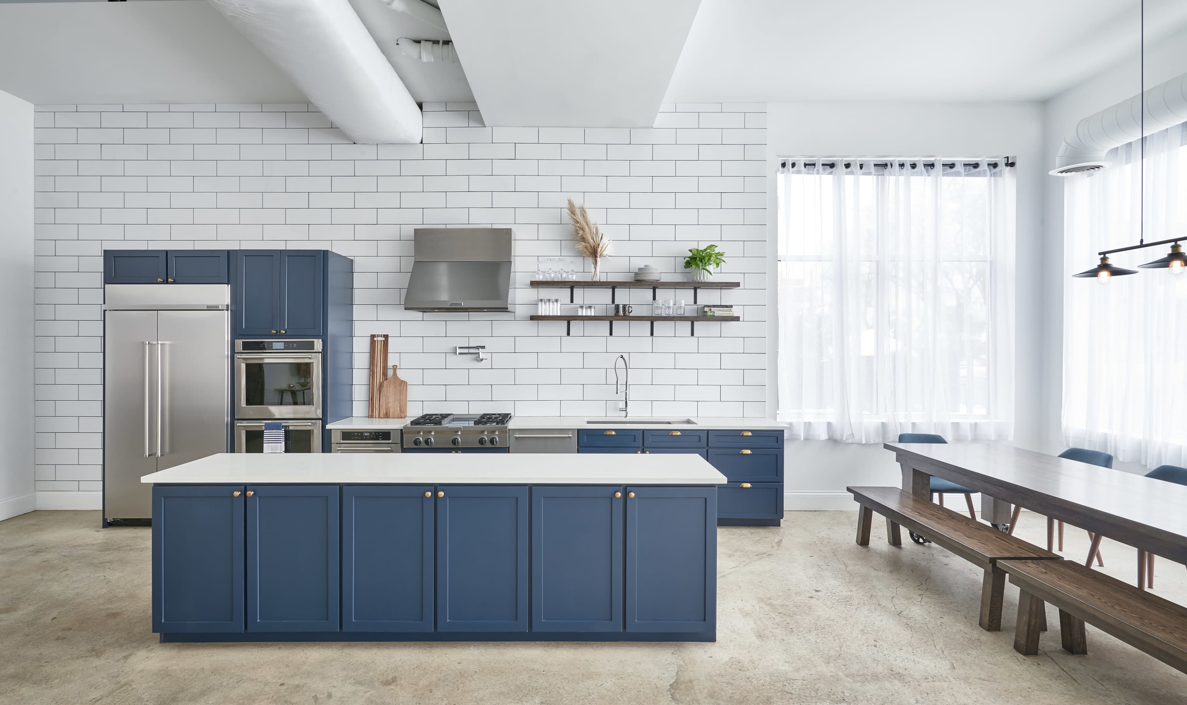The image features a modern kitchen with navy blue cabinetry, a stainless steel refrigerator, a large central island, and a dining table made of wood nearby.