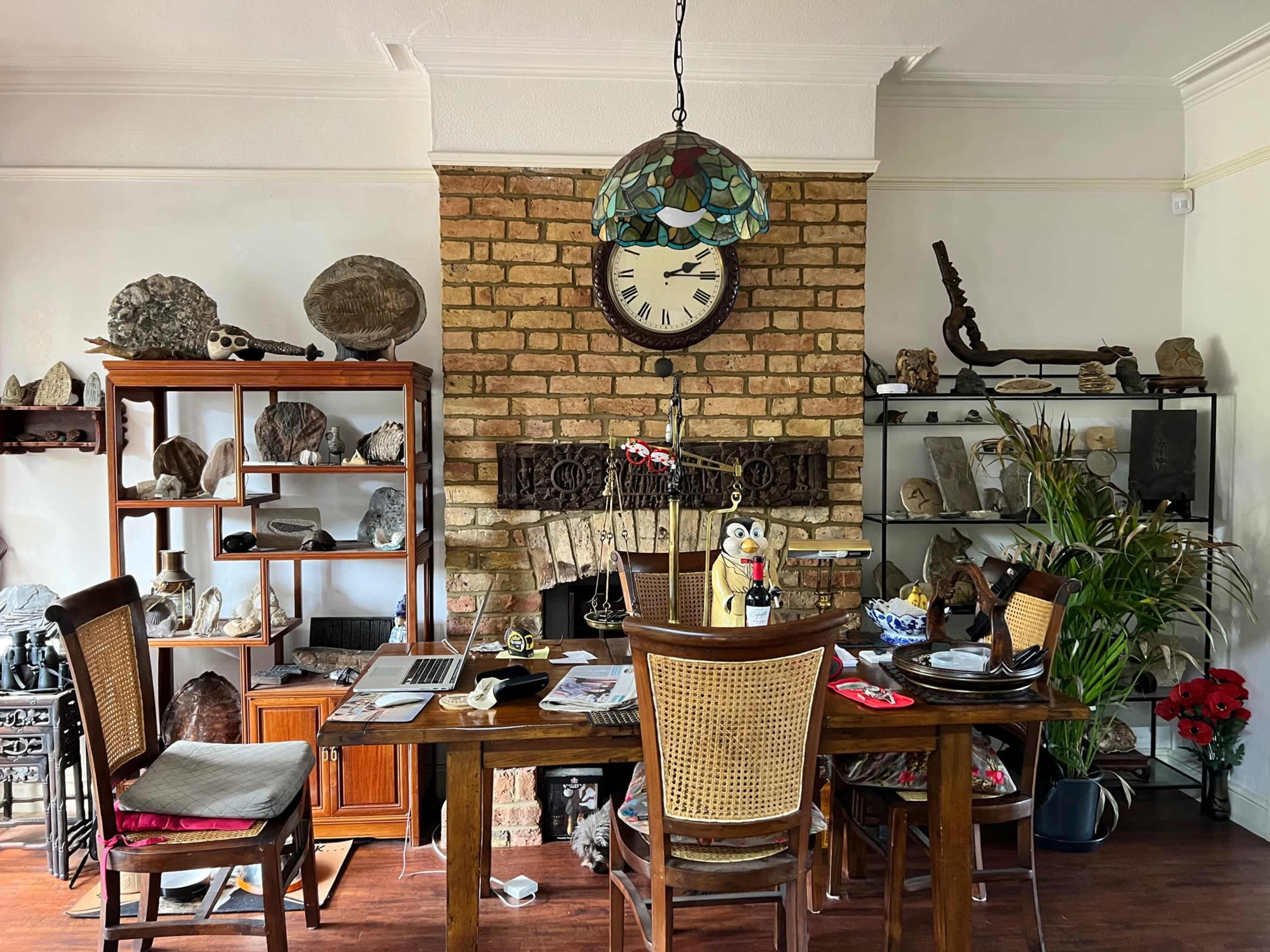 A dining area with a wooden table and chairs, a large clock on the brick wall, and shelves filled with various decorative rocks and artifacts.