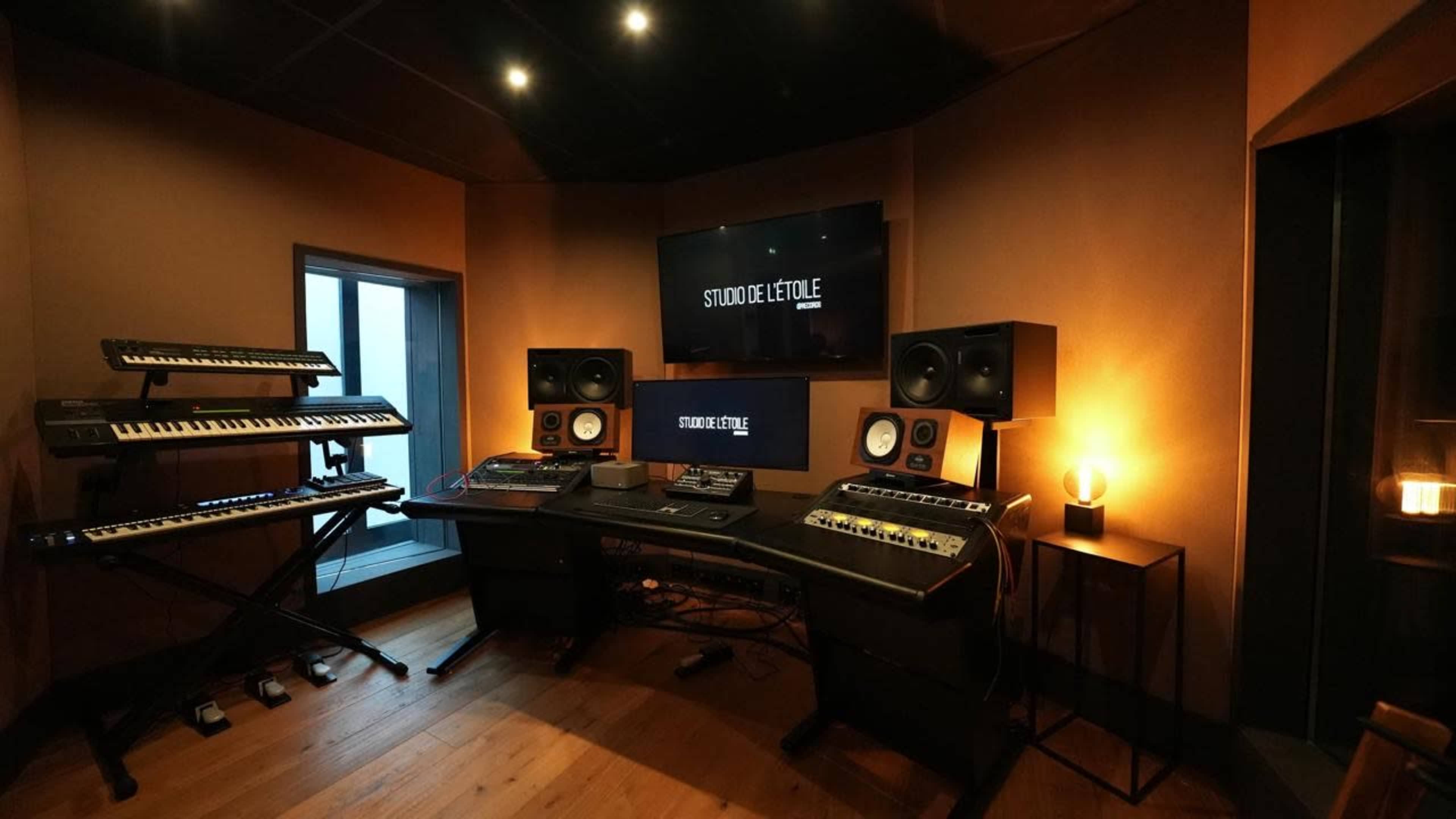 The image shows a modern music studio with keyboard instruments, audio monitors, a mixing console, and a digital display on the wall.