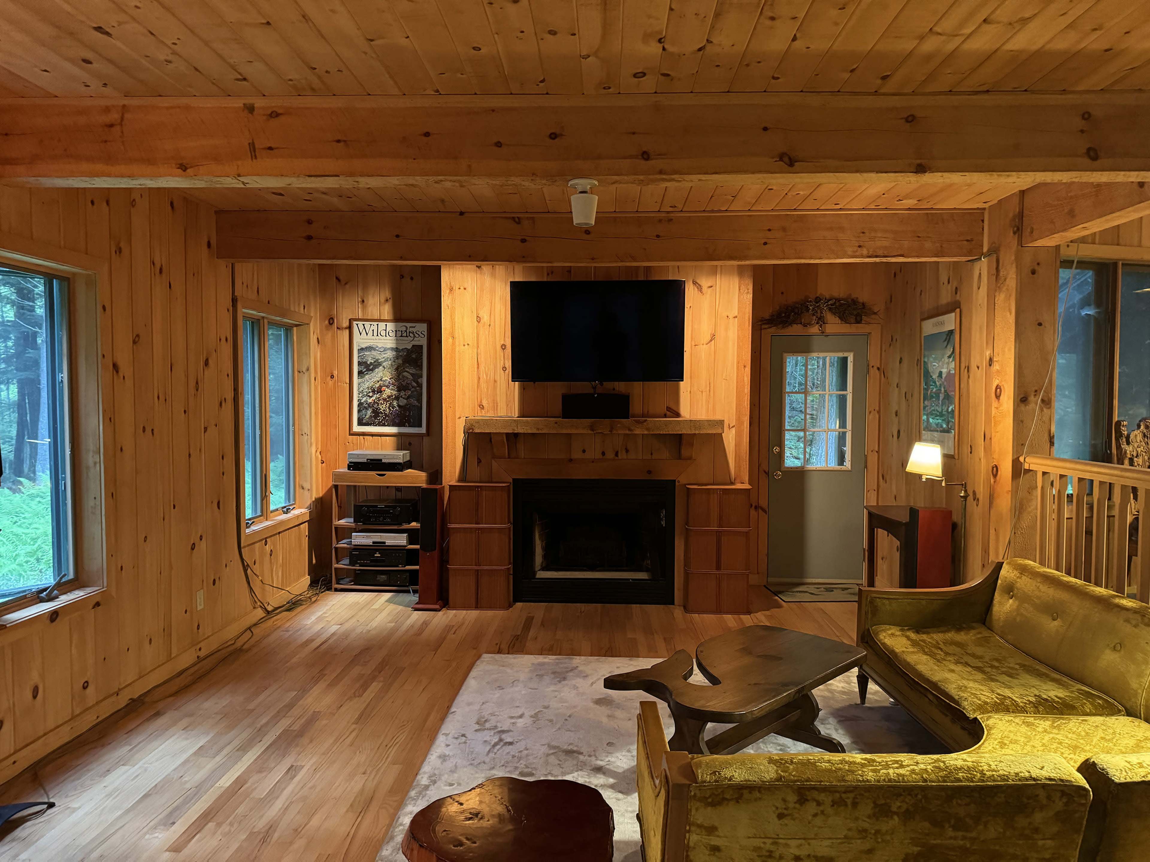 The interior of a wooden cabin features a cozy living area with a fireplace, a television mounted on the wall, and a yellow couch facing a coffee table.