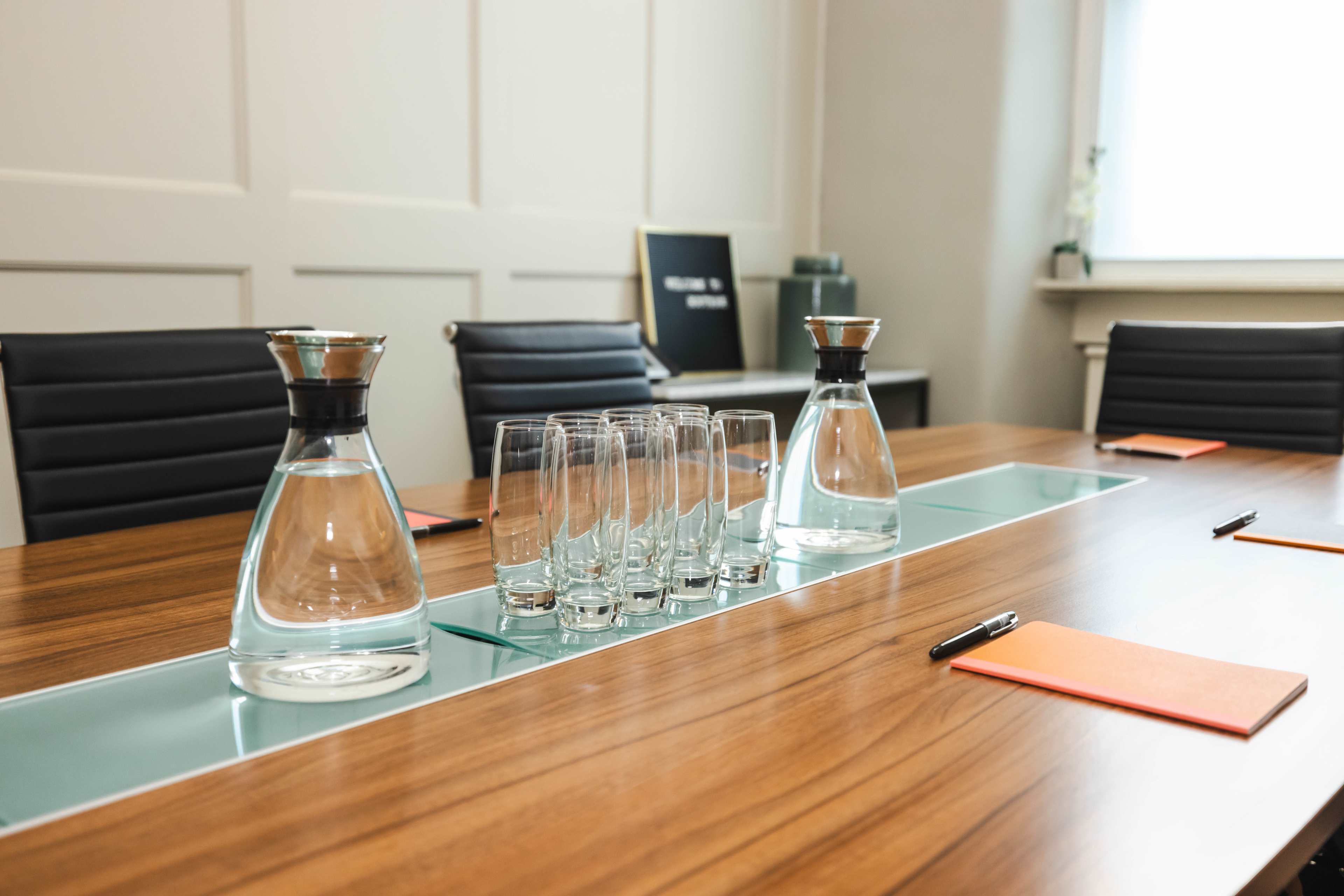 A wooden conference table is set with glass water carafes, empty glasses, and orange notepads beside black chairs.
