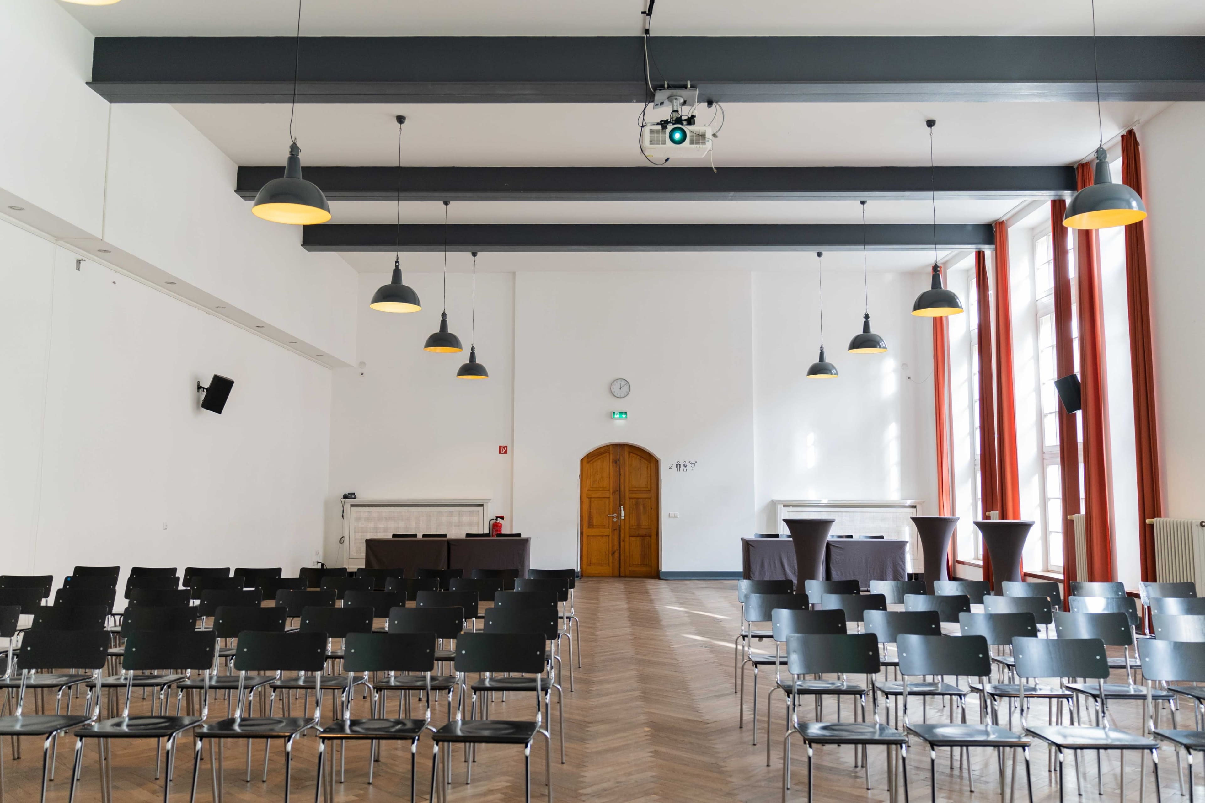 A spacious meeting room with rows of black chairs, high ceilings, and large windows, featuring a projector mounted on the ceiling.