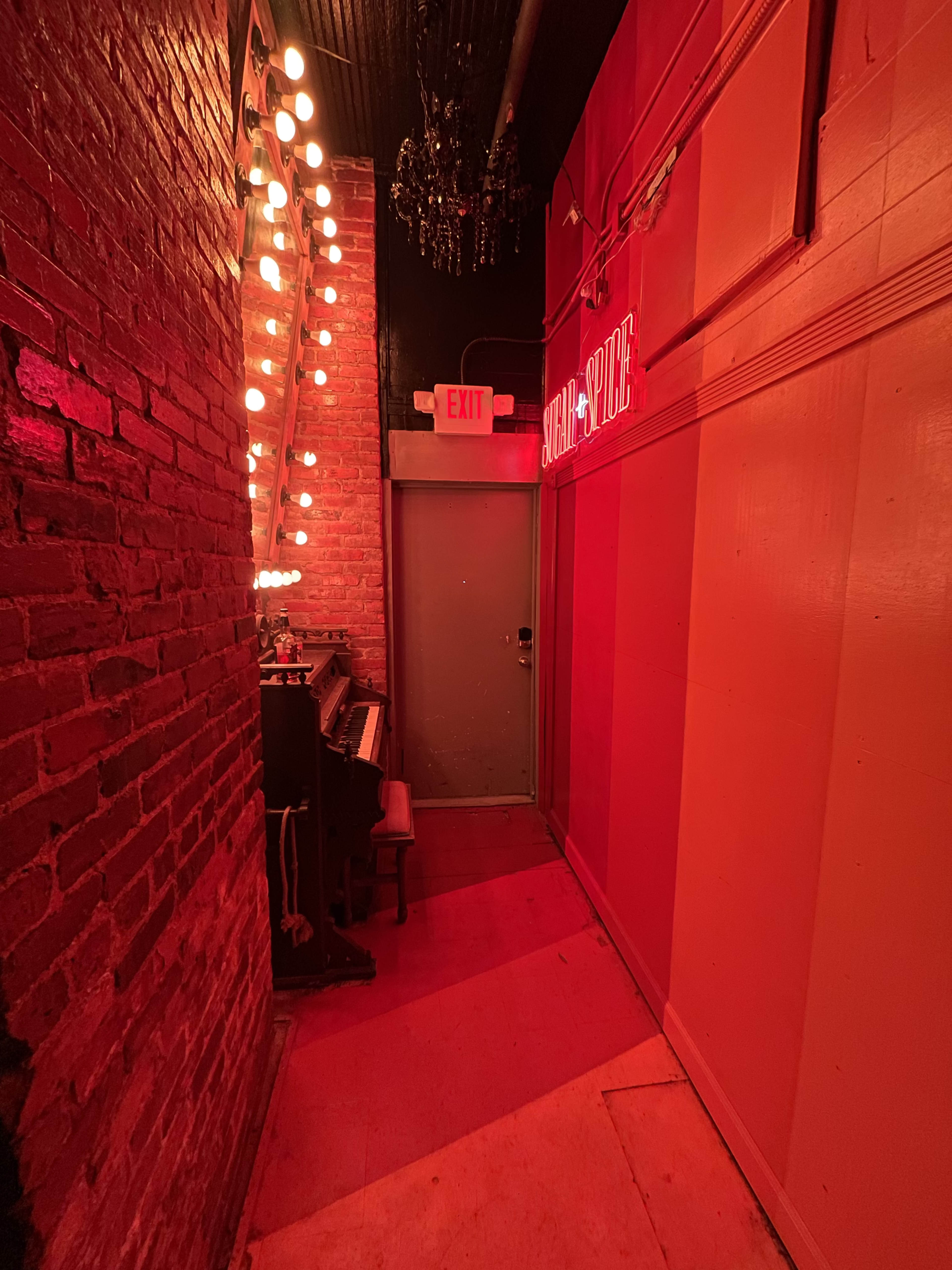 A narrow hallway features exposed brick walls, dim red lighting, and a vintage piano next to a door marked with an exit sign.