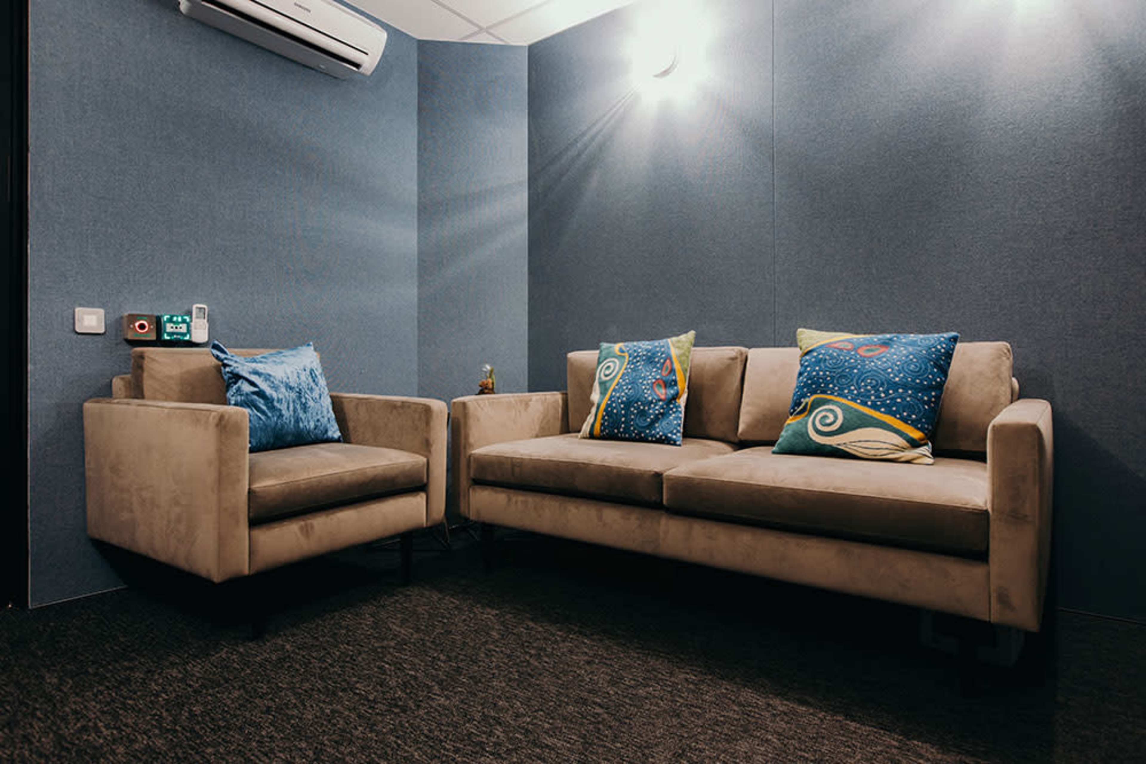 A small, modern lounge area with a beige sofa and an armchair, both adorned with colorful patterned cushions, set against blue walls and carpet.