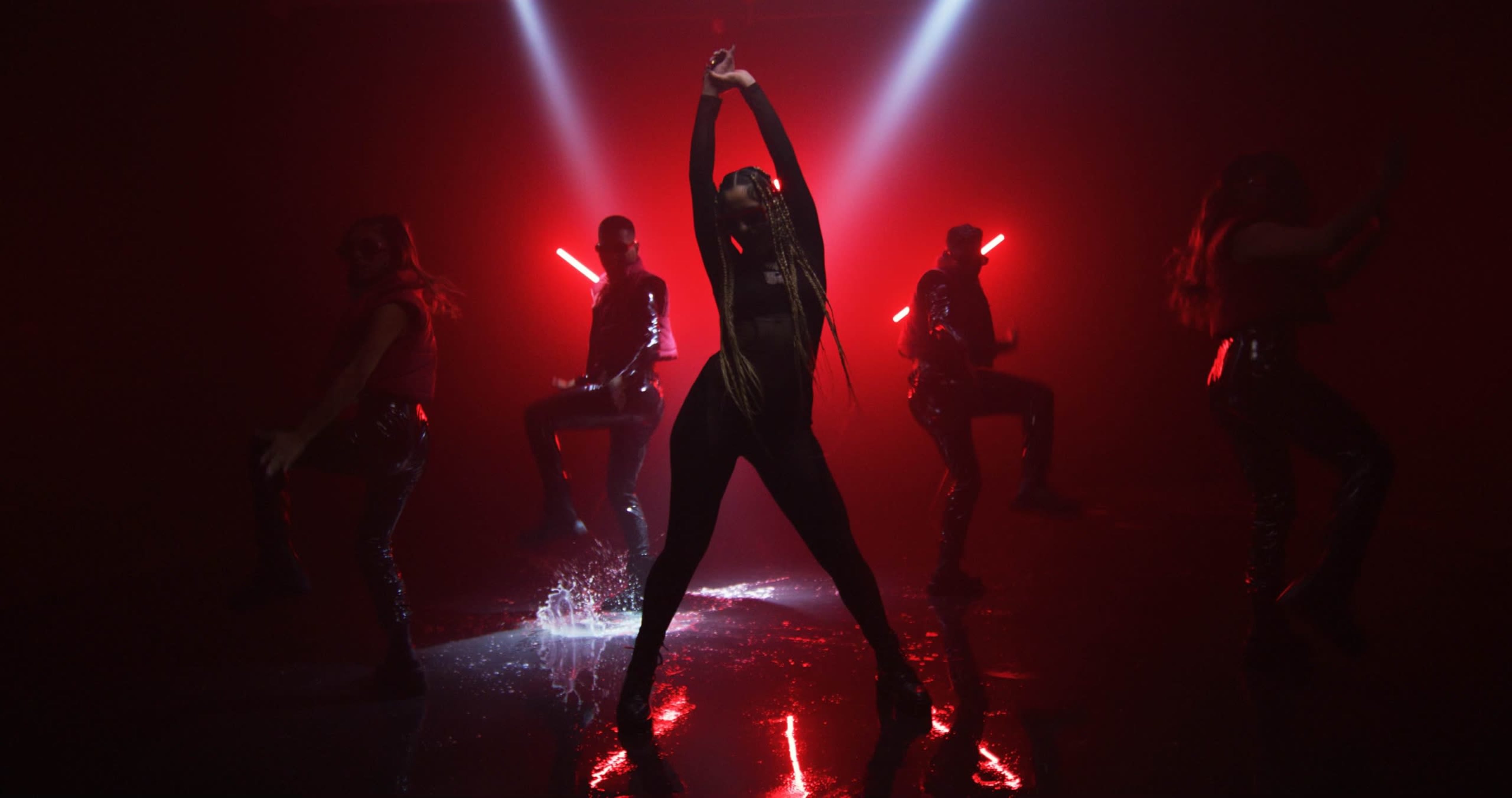 A dancer strikes a pose in the spotlight while three backup performers move in the background amidst dramatic lighting and reflections on a wet surface.