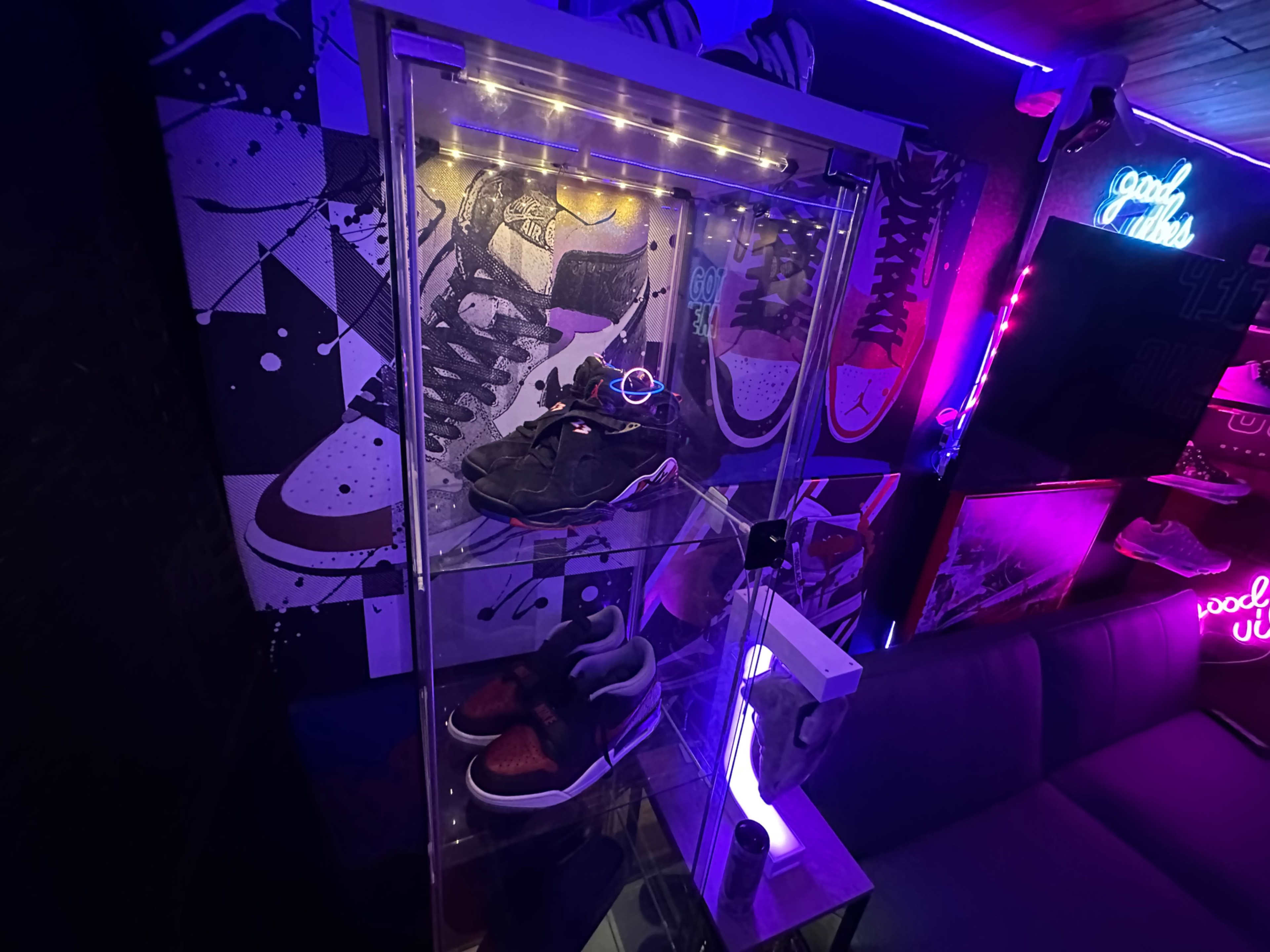 The image shows a display case illuminated with LED lights, showcasing two pairs of sneakers against a colorful, sneaker-themed wall mural.