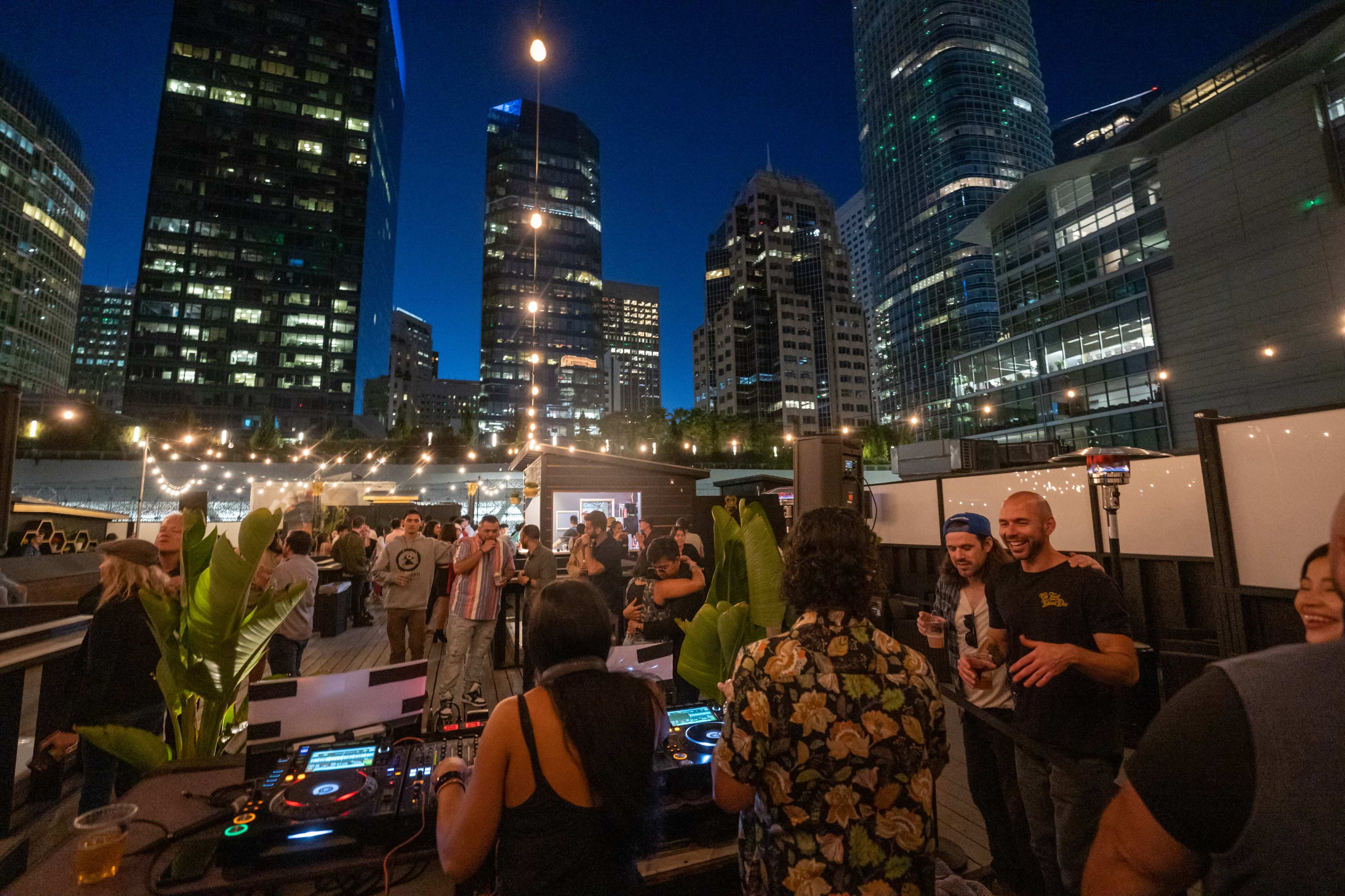A rooftop party is taking place at night with people dancing and socializing, while a DJ plays music in front of tall city buildings.