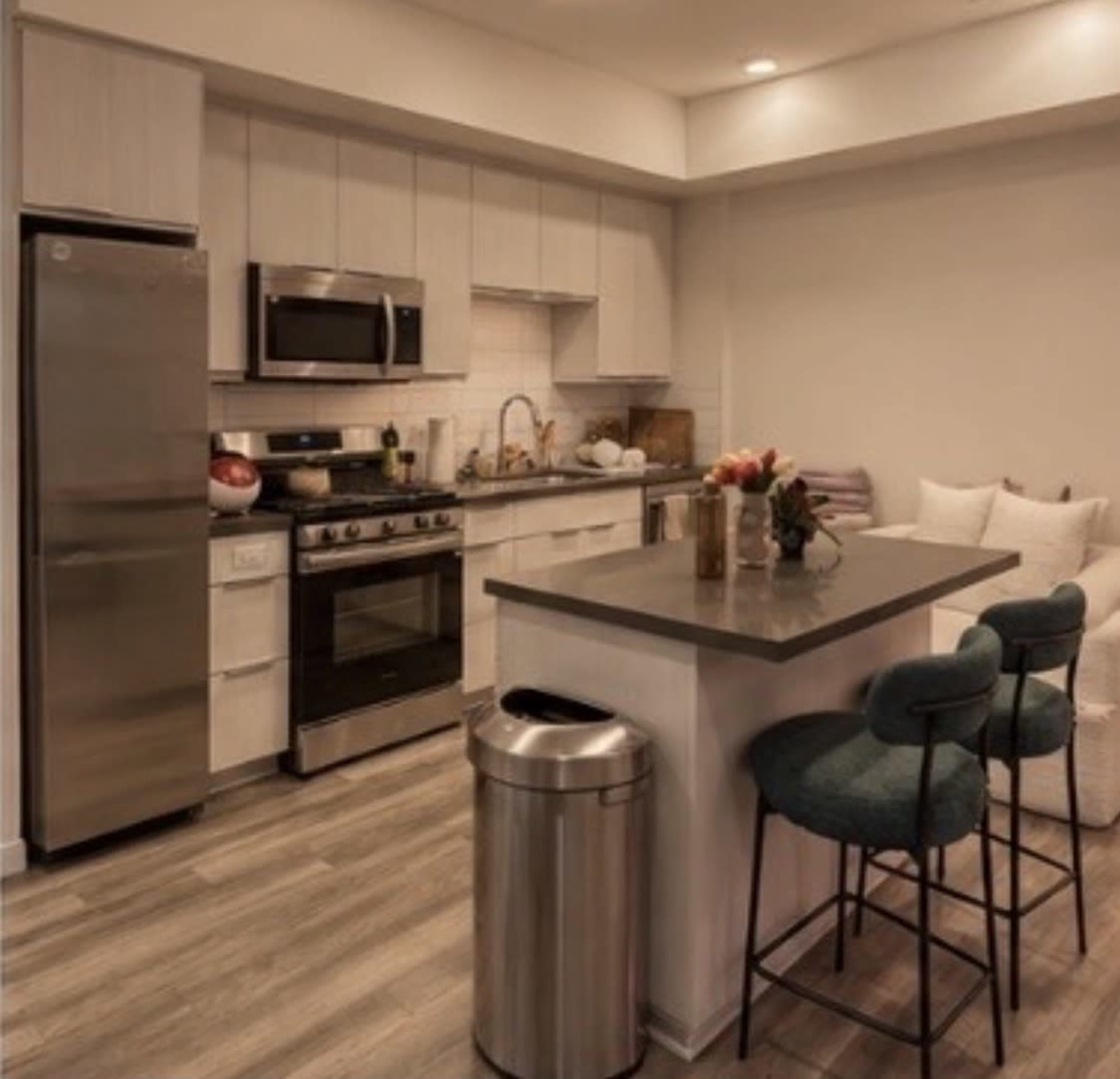 The image shows a modern kitchen with stainless steel appliances, a central island with bar stools, and a small dining area.