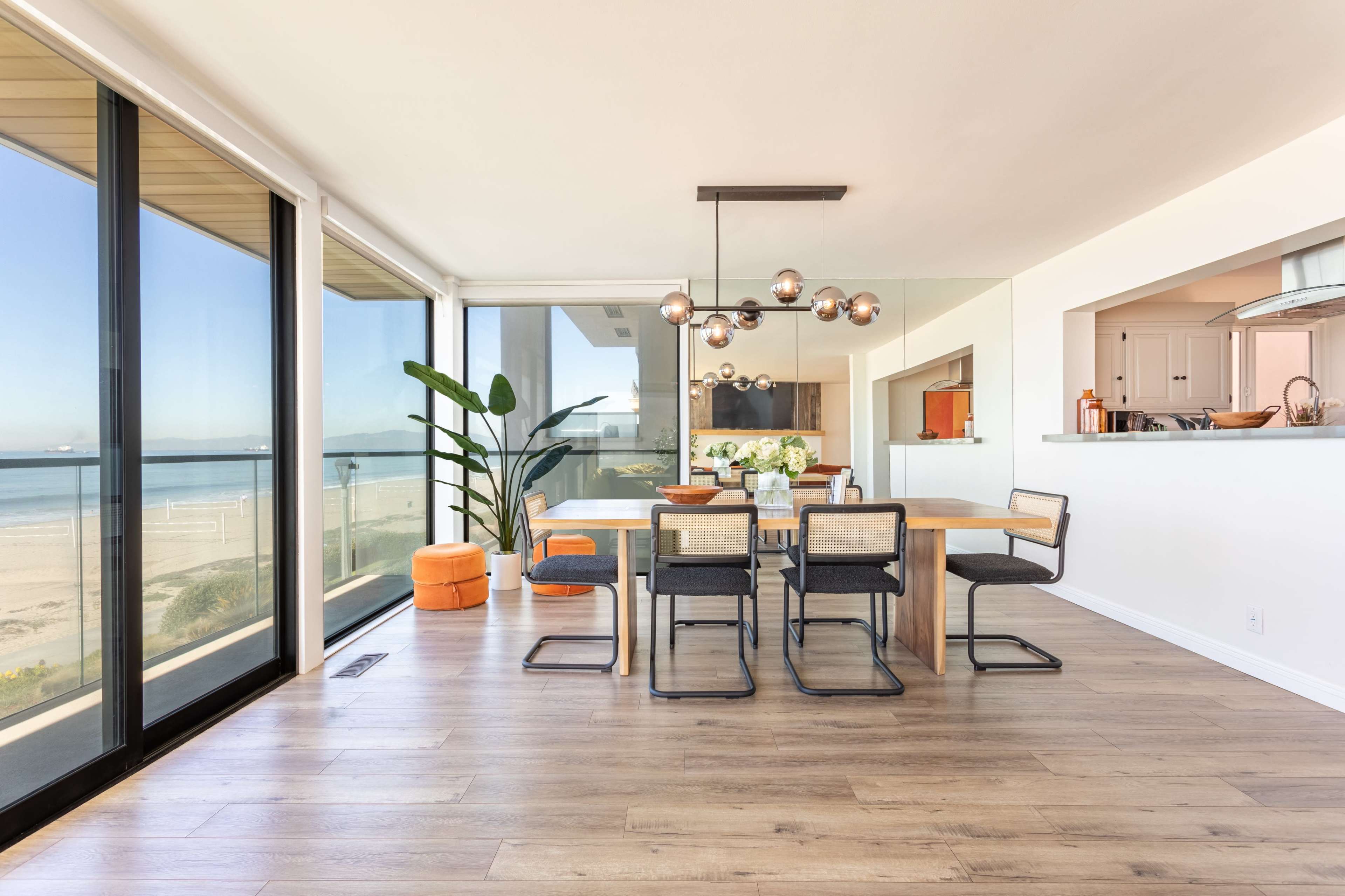 A dining area features a wooden table surrounded by black chairs, large windows showcasing a beach view, and modern light fixtures hanging overhead.