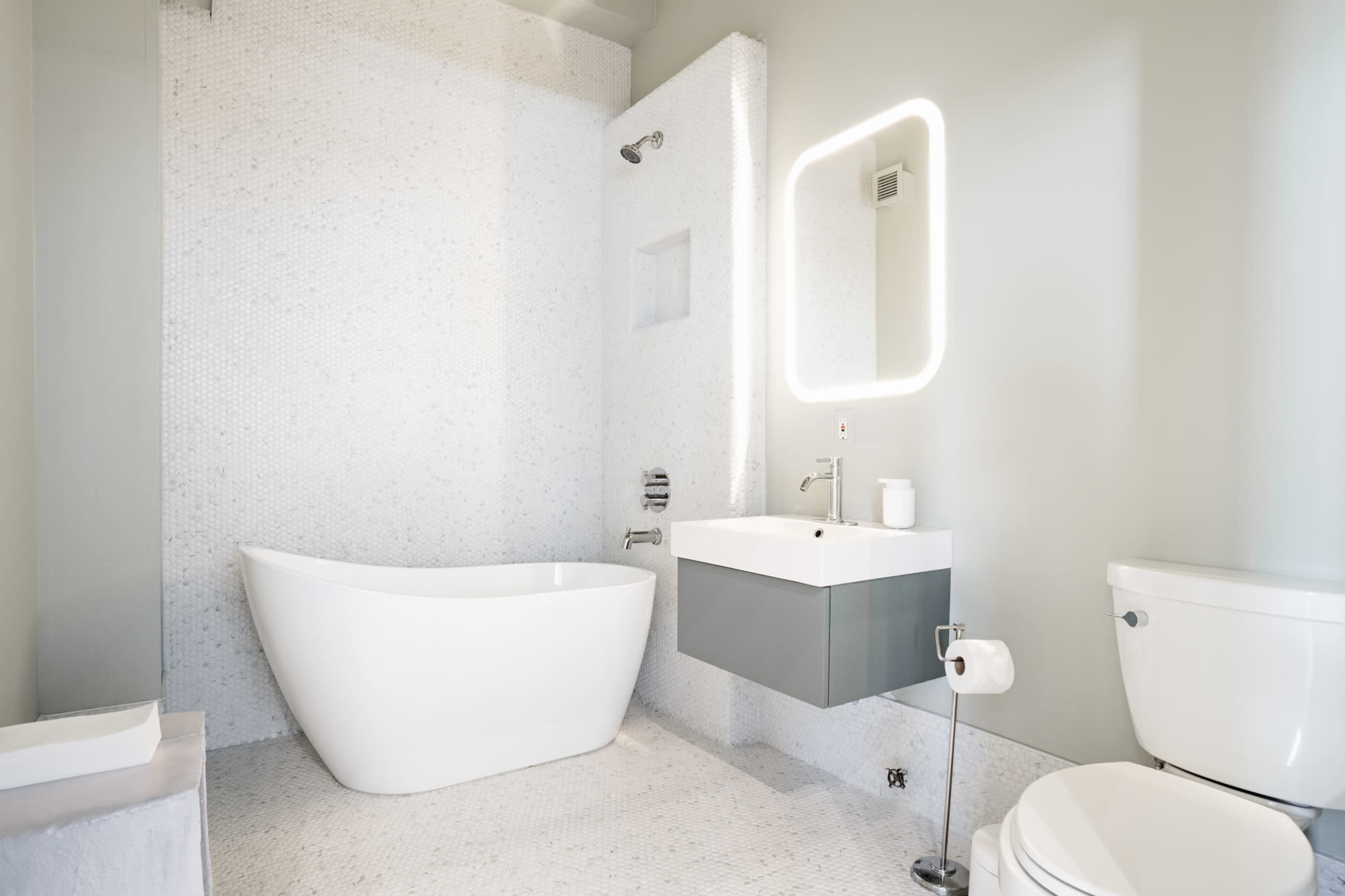 The image shows a modern bathroom featuring a freestanding bathtub, a wall-mounted sink with a mirror, a toilet, and pebble tile flooring.