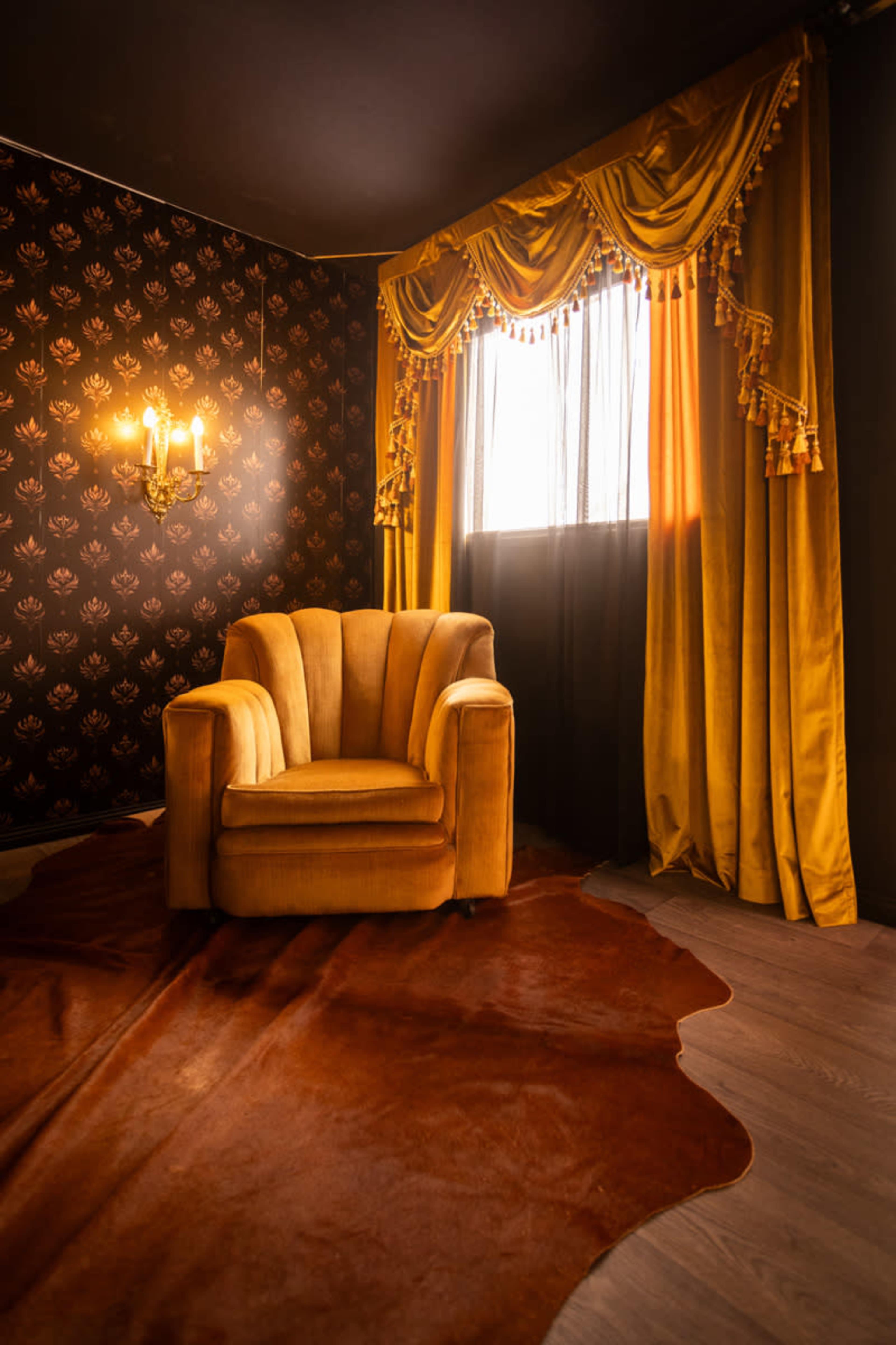 The image shows a plush yellow armchair positioned next to a window with golden curtains, set against a dark floral-patterned wallpaper and a brown area rug.