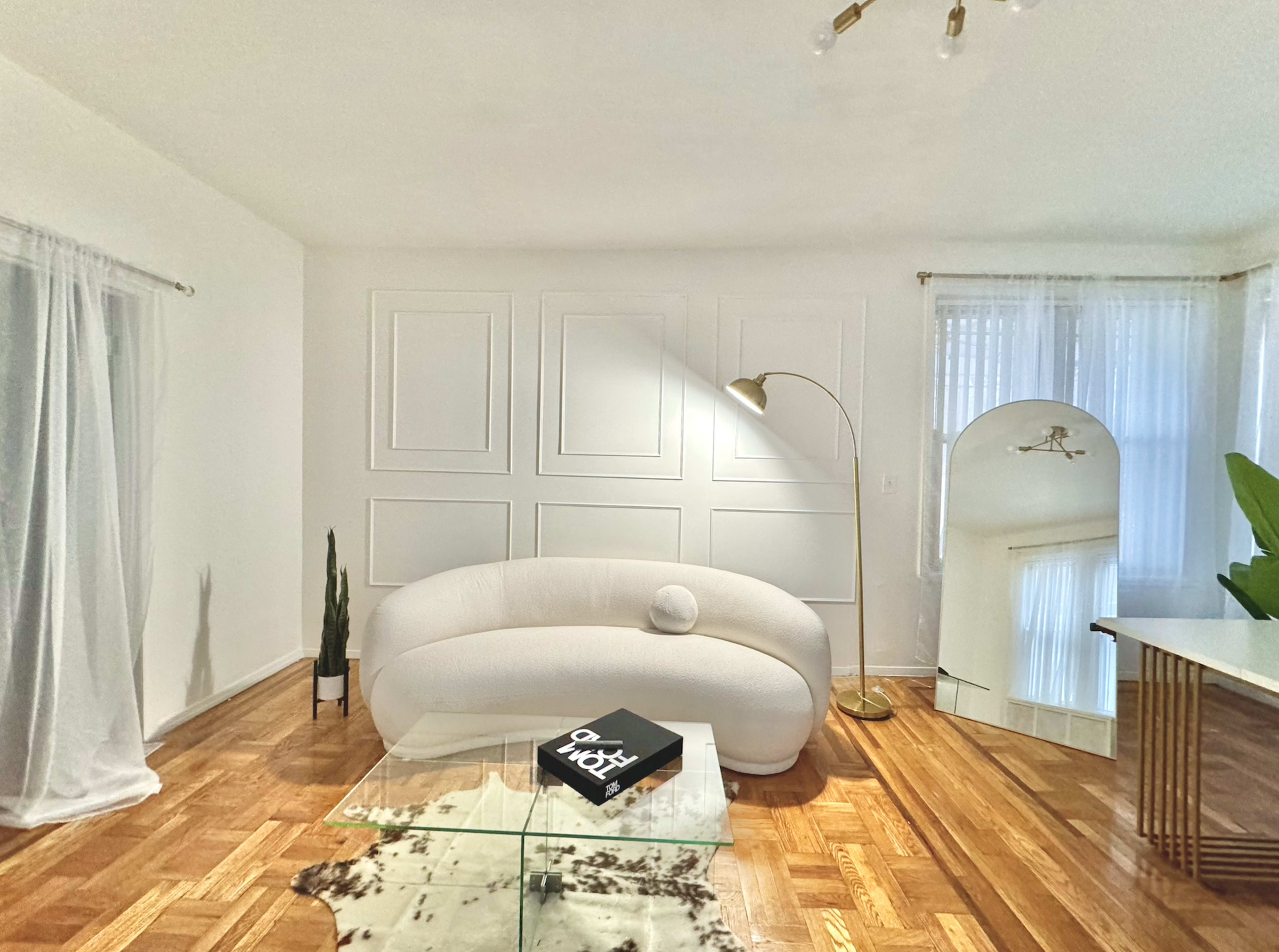 A modern living room featuring a white curved sofa, a floor lamp, a glass coffee table, and a tall plant, with decorative wall panels and large windows allowing natural light.