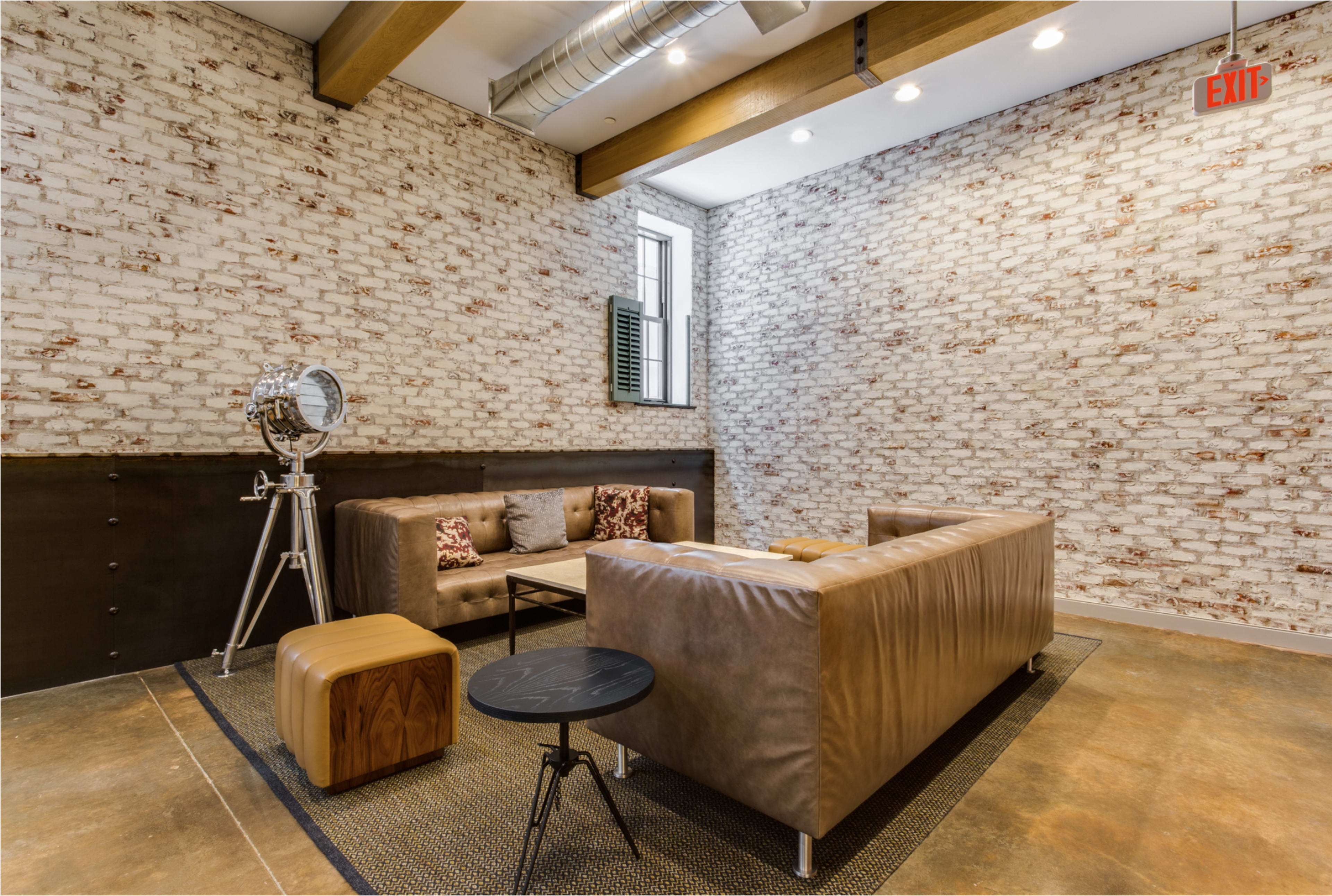 The image features a modern seating area with a leather couch, a small wooden table, and a vintage-style floor lamp against a brick wall.