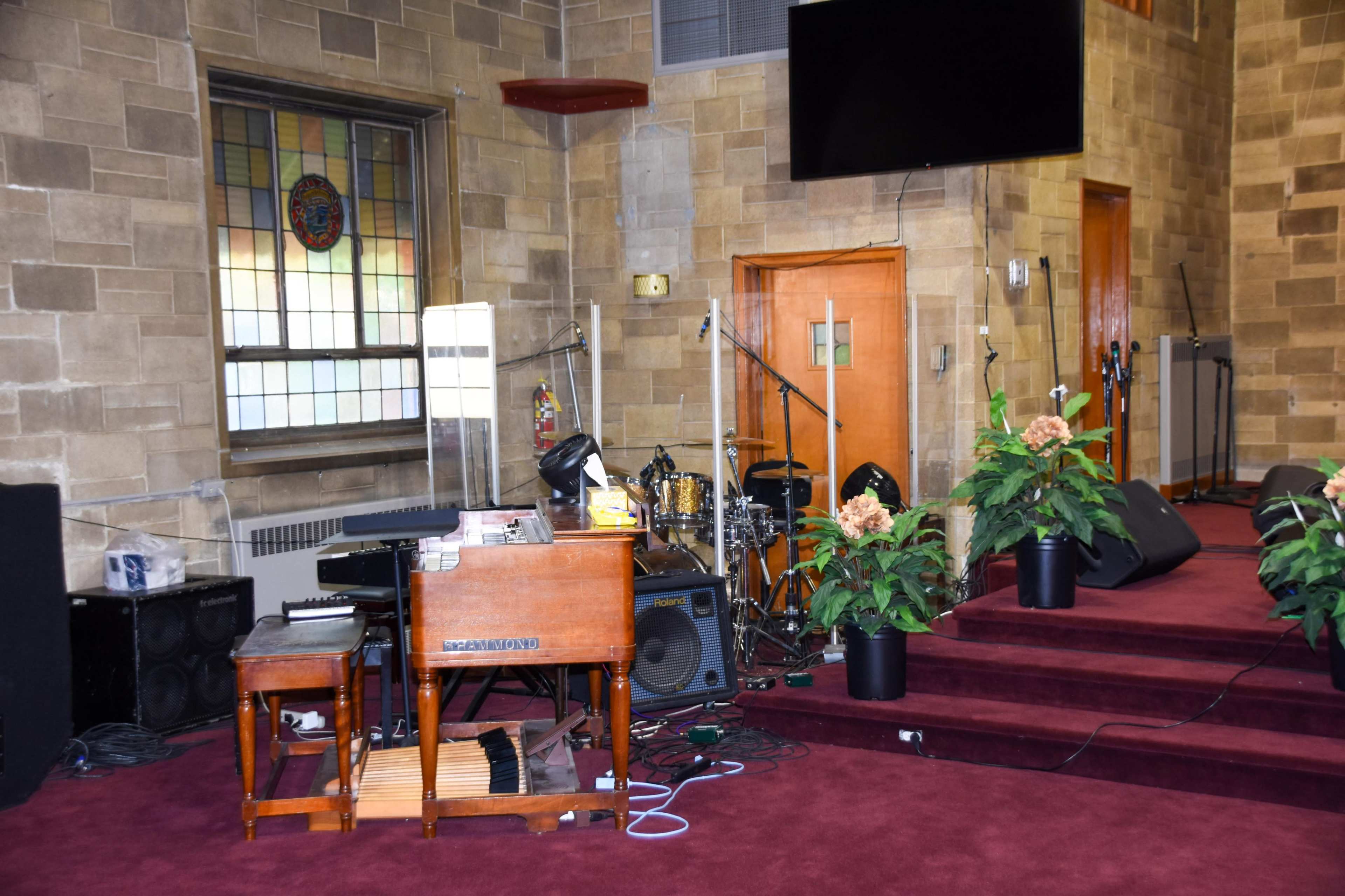 The image shows a small stage setup in a church with musical instruments, including a piano and drums, surrounded by potted plants and a large screen on the wall.