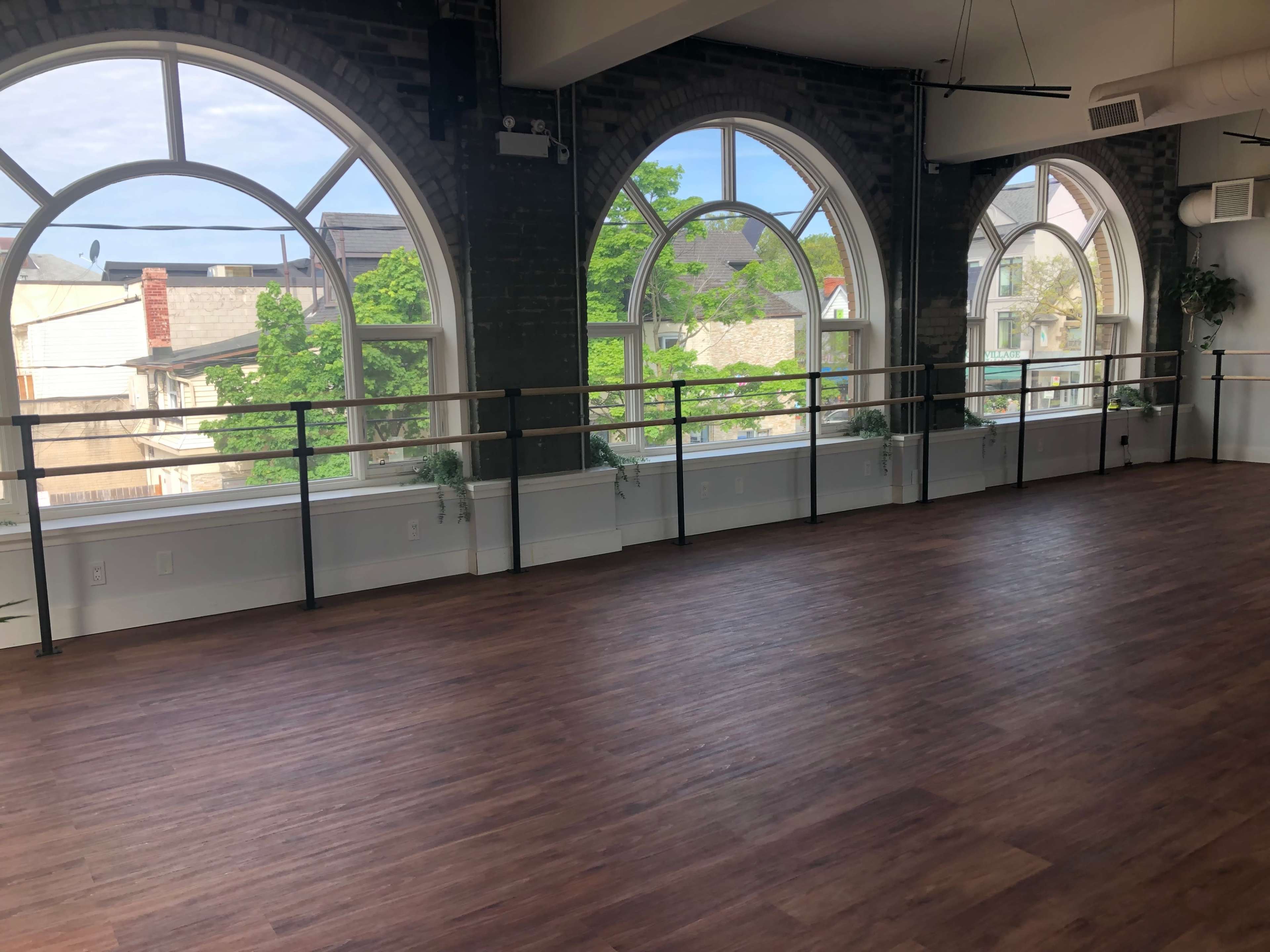 The image shows a spacious dance studio with large arched windows overlooking a tree-lined street.