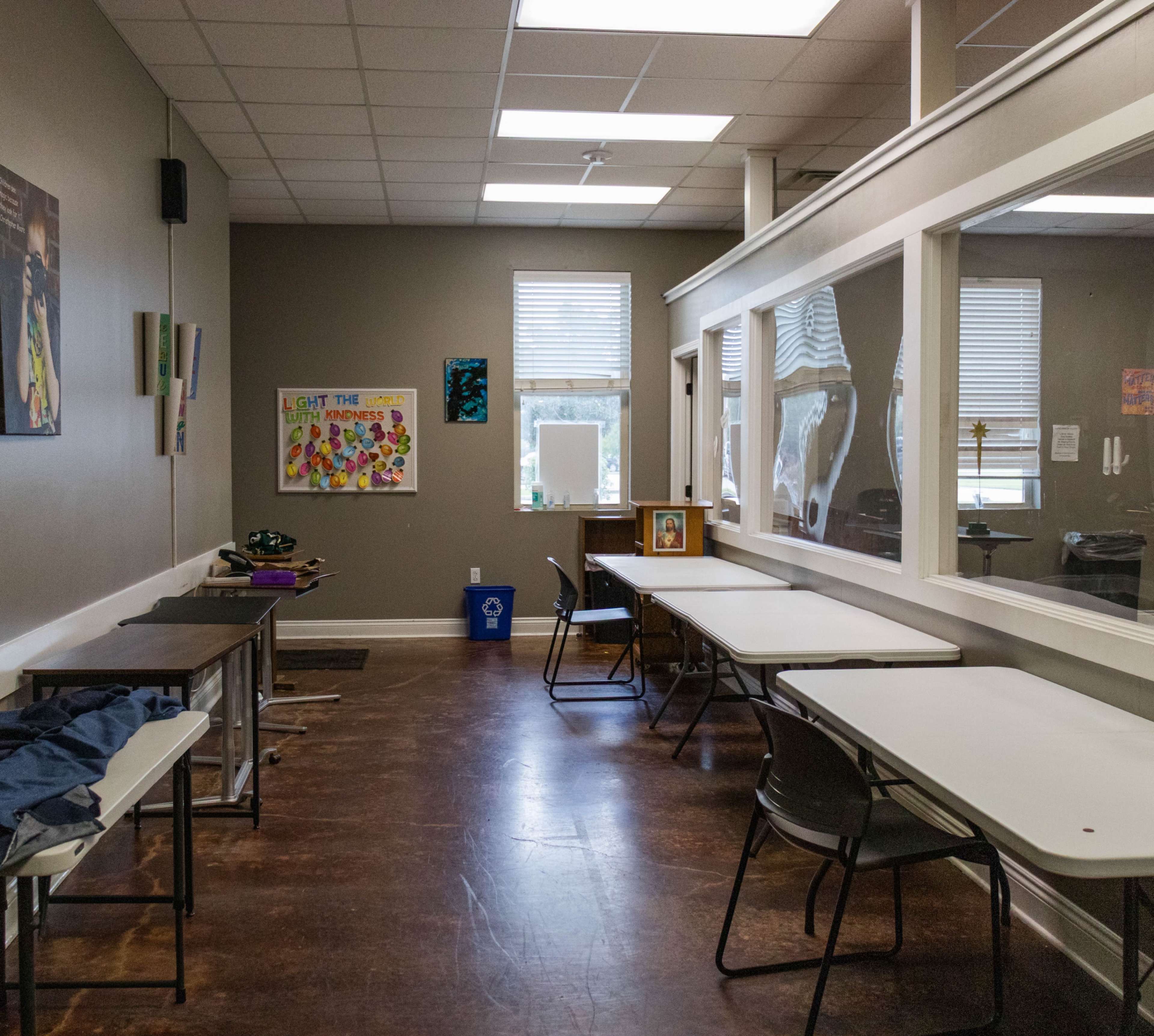 The image displays a classroom with several tables and chairs arranged along the walls, with windows letting in natural light and artwork hanging on the walls.