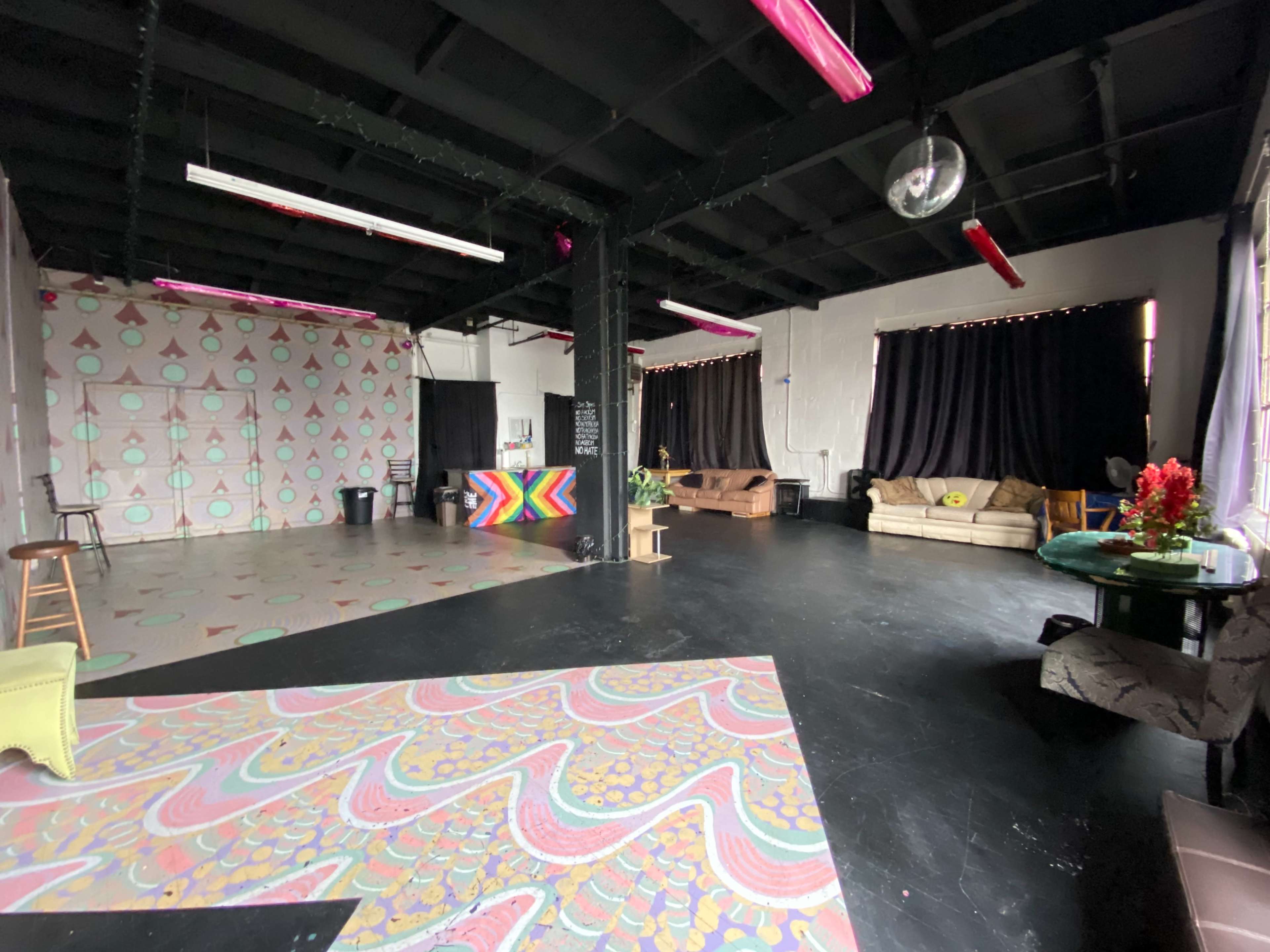 The image shows a spacious studio with a patterned floor, colorful wall art, and various seating arrangements, including couches and stools.
