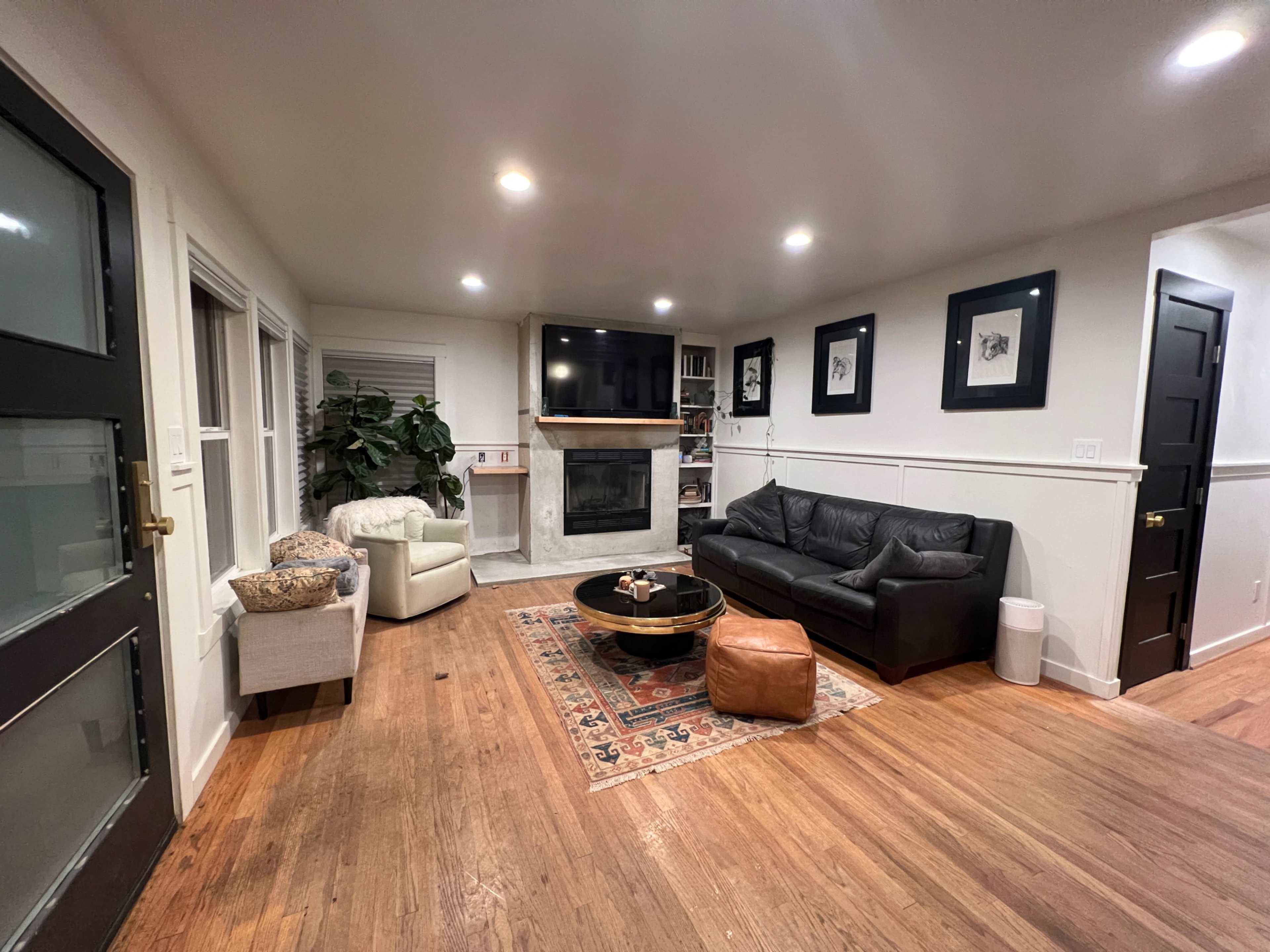 The living room features a leather sofa, a round coffee table, a flat-screen TV above a fireplace, and light wood flooring with a decorative rug.
