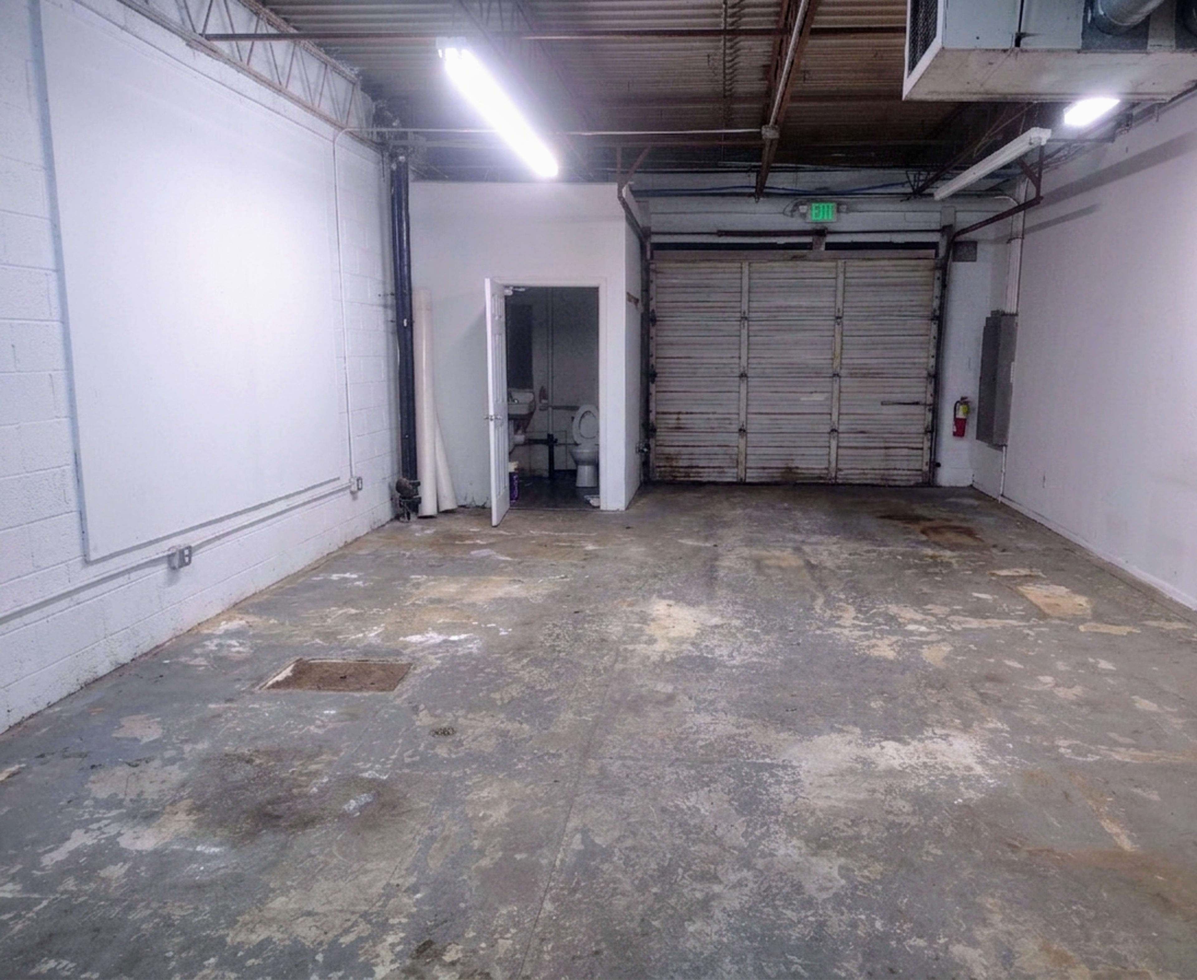 The image shows an empty warehouse space with concrete flooring, white walls, and a partially open garage door at the far end.