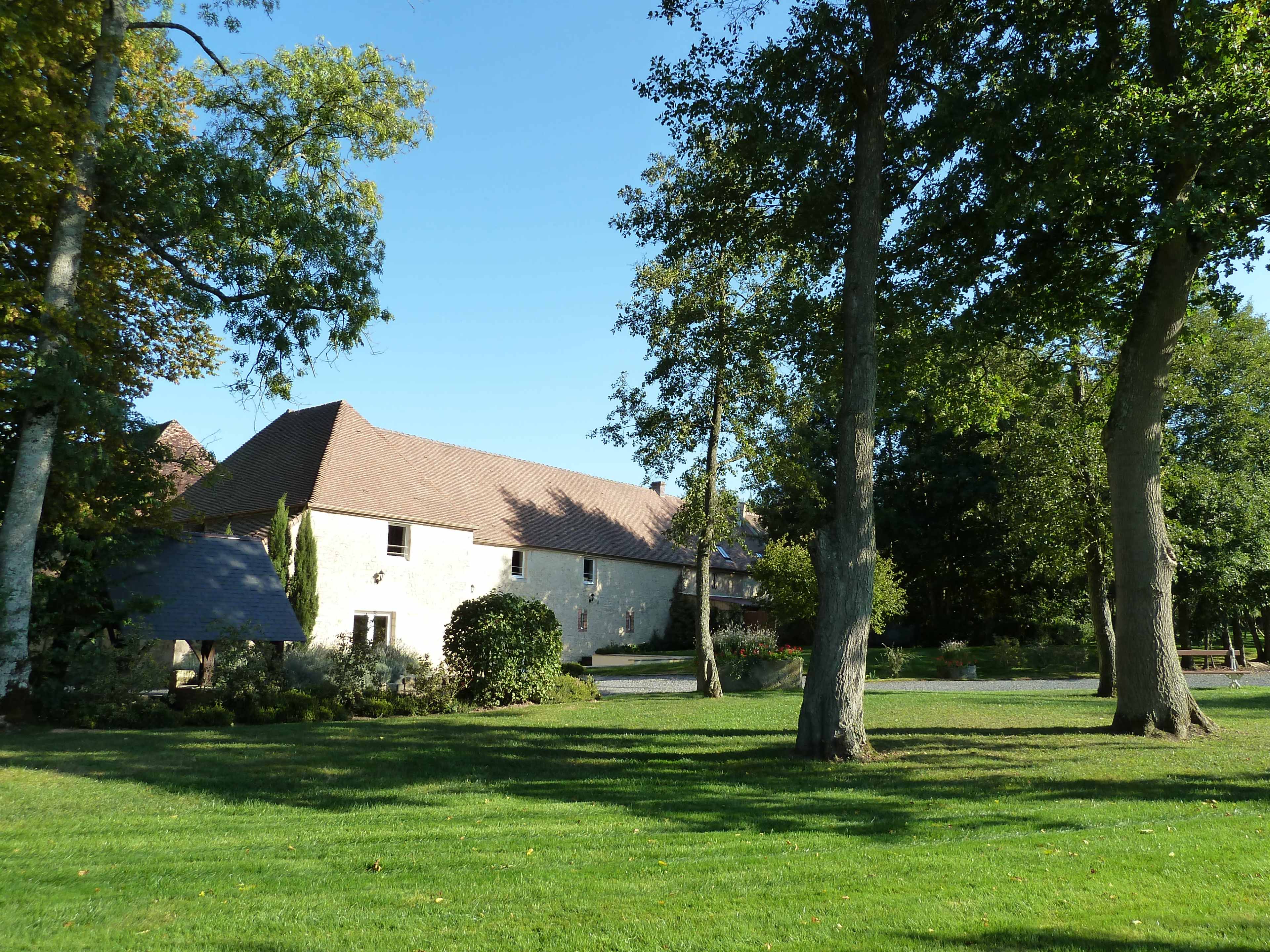 A large, white stone building with a brown roof is surrounded by green grass and tall trees under a clear blue sky.