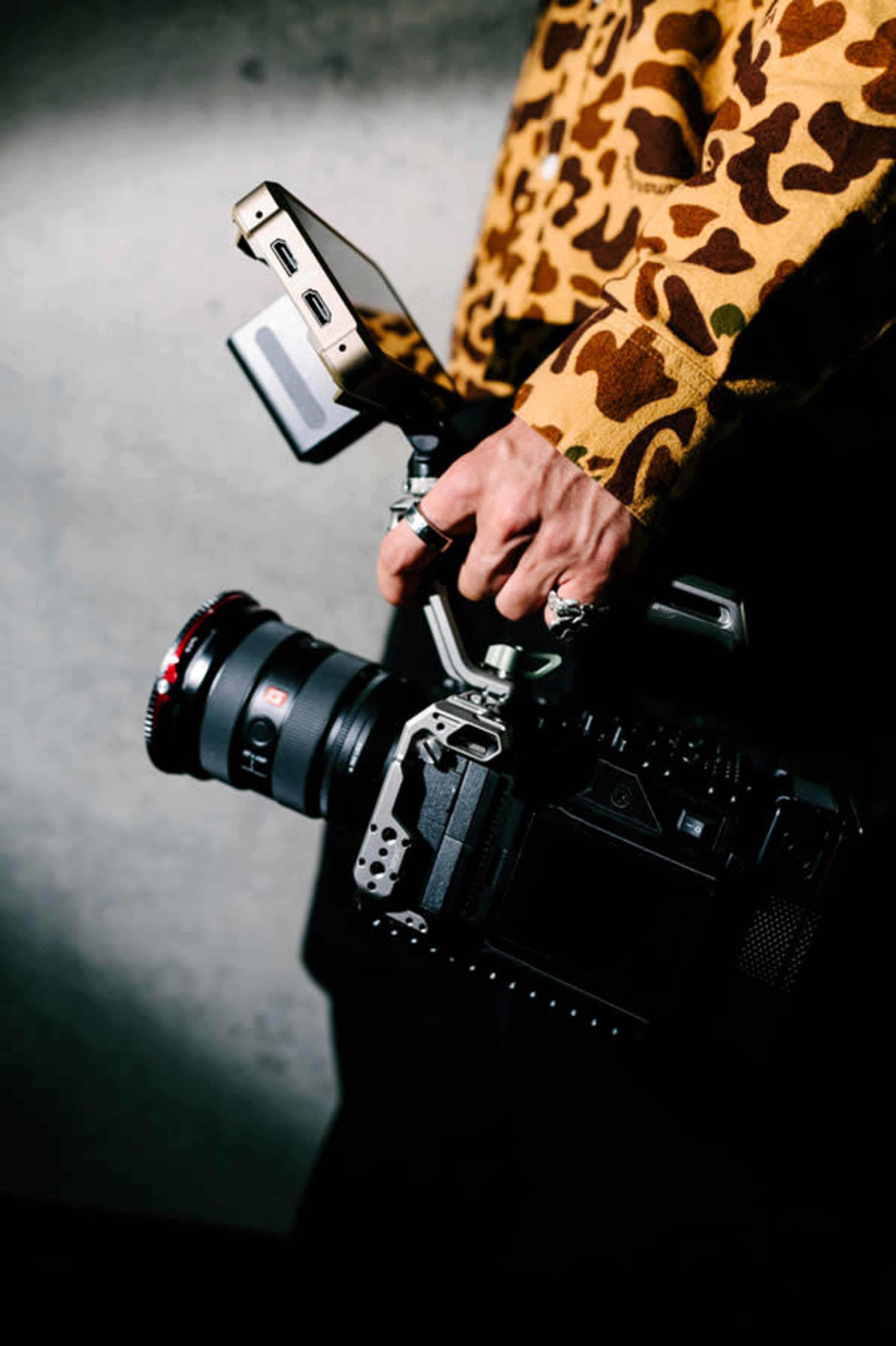 A person in a patterned shirt holds a professional video camera with a side display and a large lens.