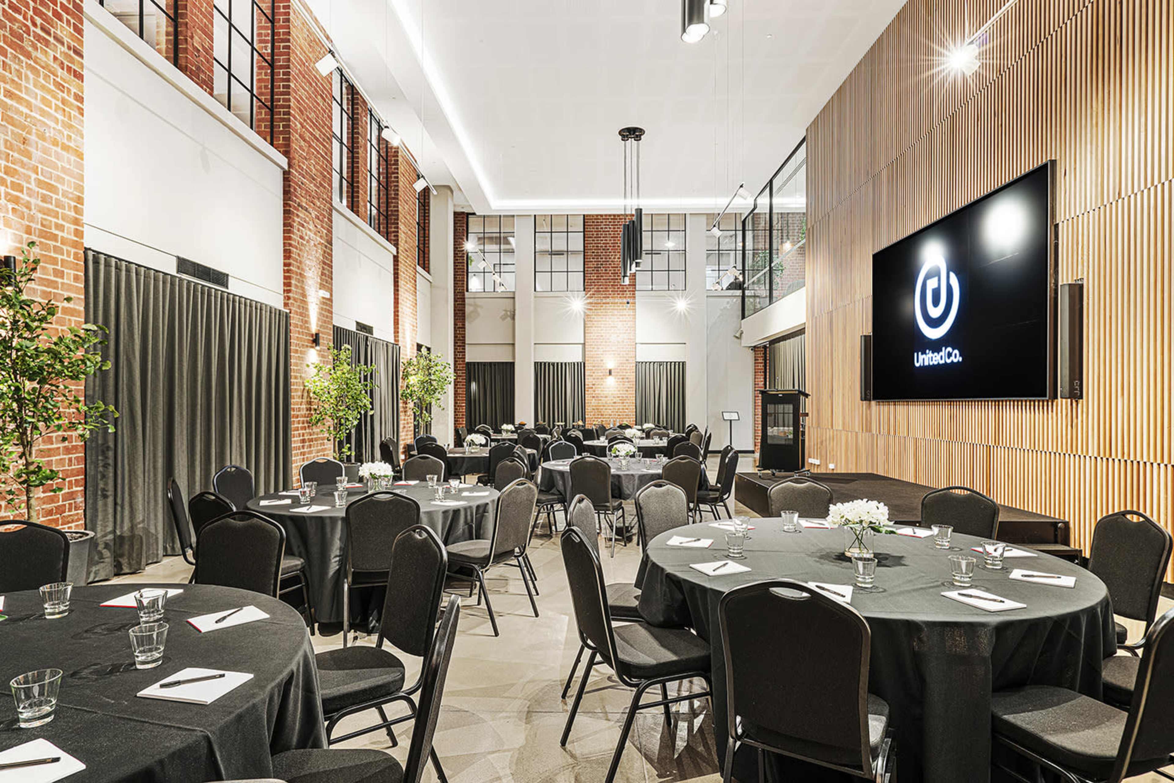 The image shows a spacious indoor event venue with round tables set for dining, featuring brick walls and modern décor.