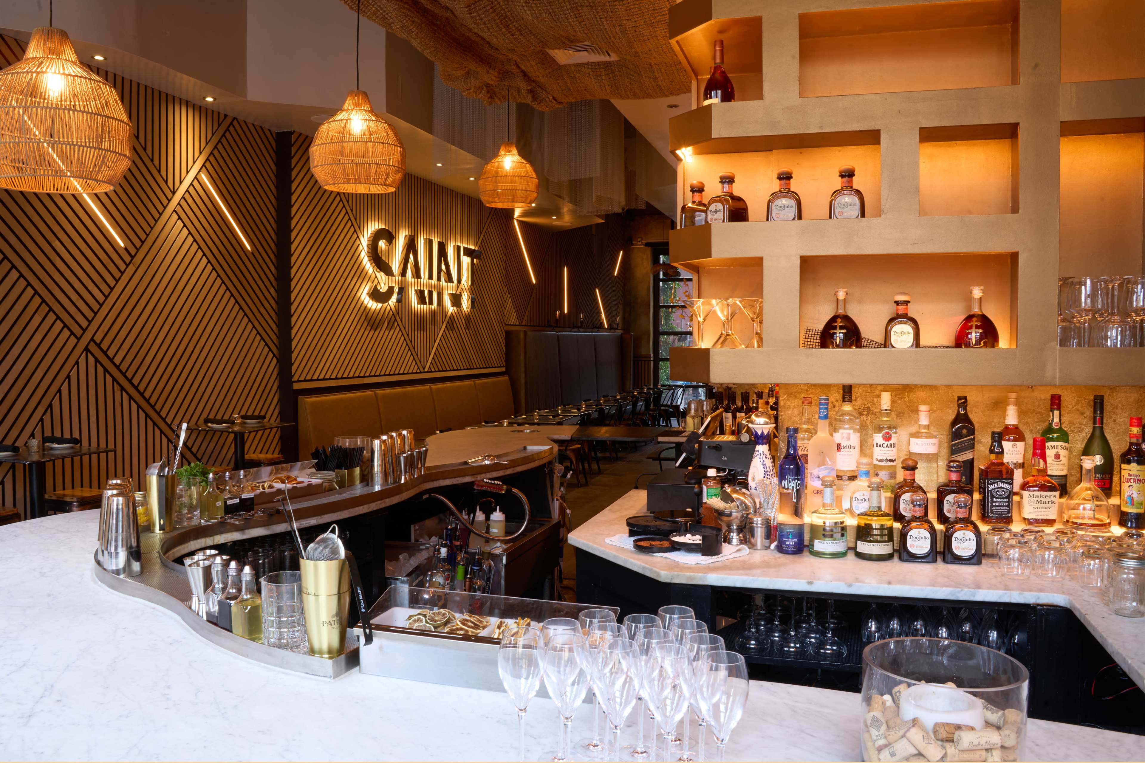 The image shows a modern bar area with a well-lit counter, an array of liquor bottles on shelves, and a geometric wooden design on the walls.