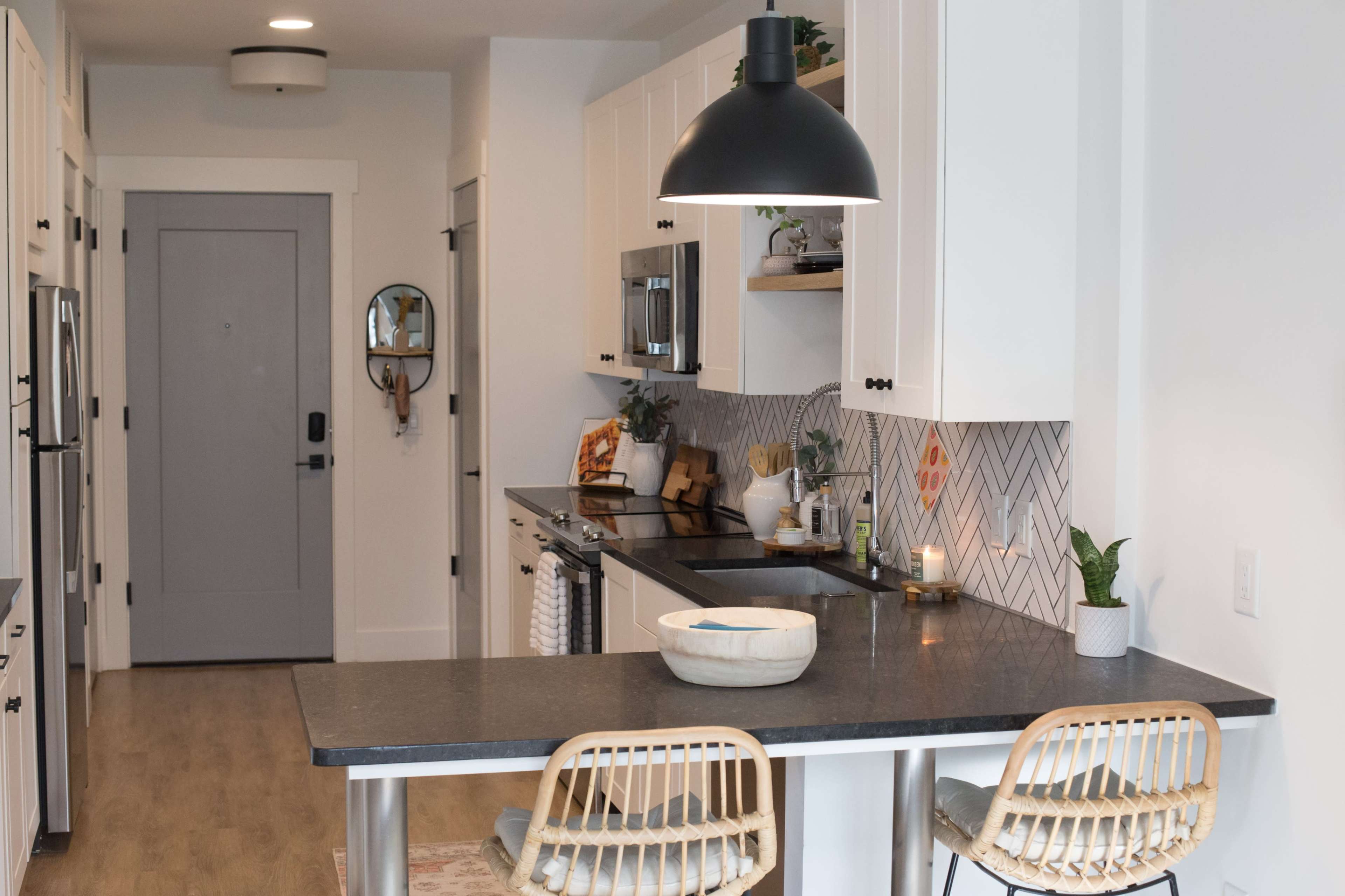 A modern kitchen features white cabinets, a black countertop, and two rattan bar stools at an island, with an open door visible in the background.