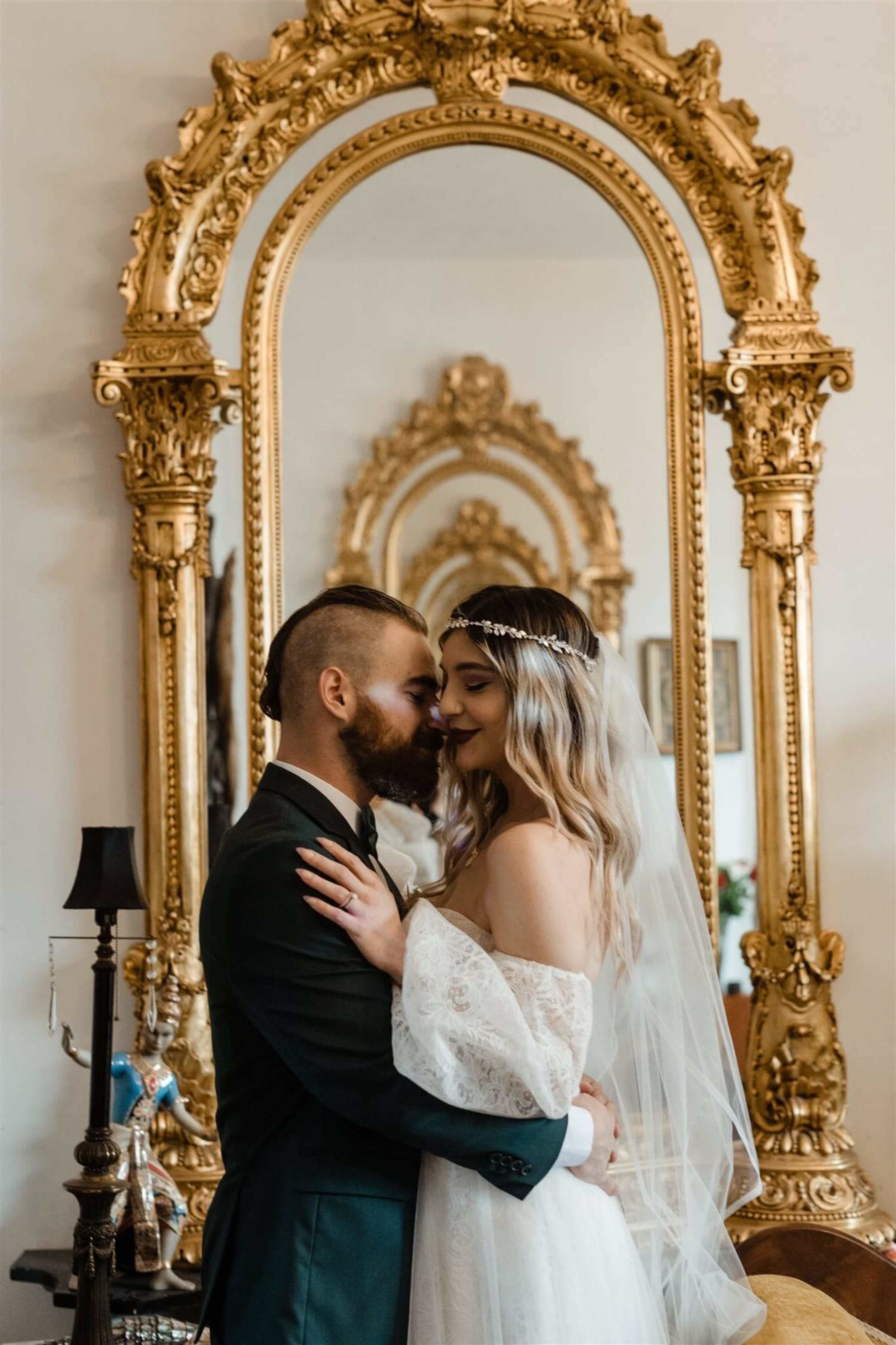 A couple embraces in front of an ornate, gilded mirror in a decorated interior.