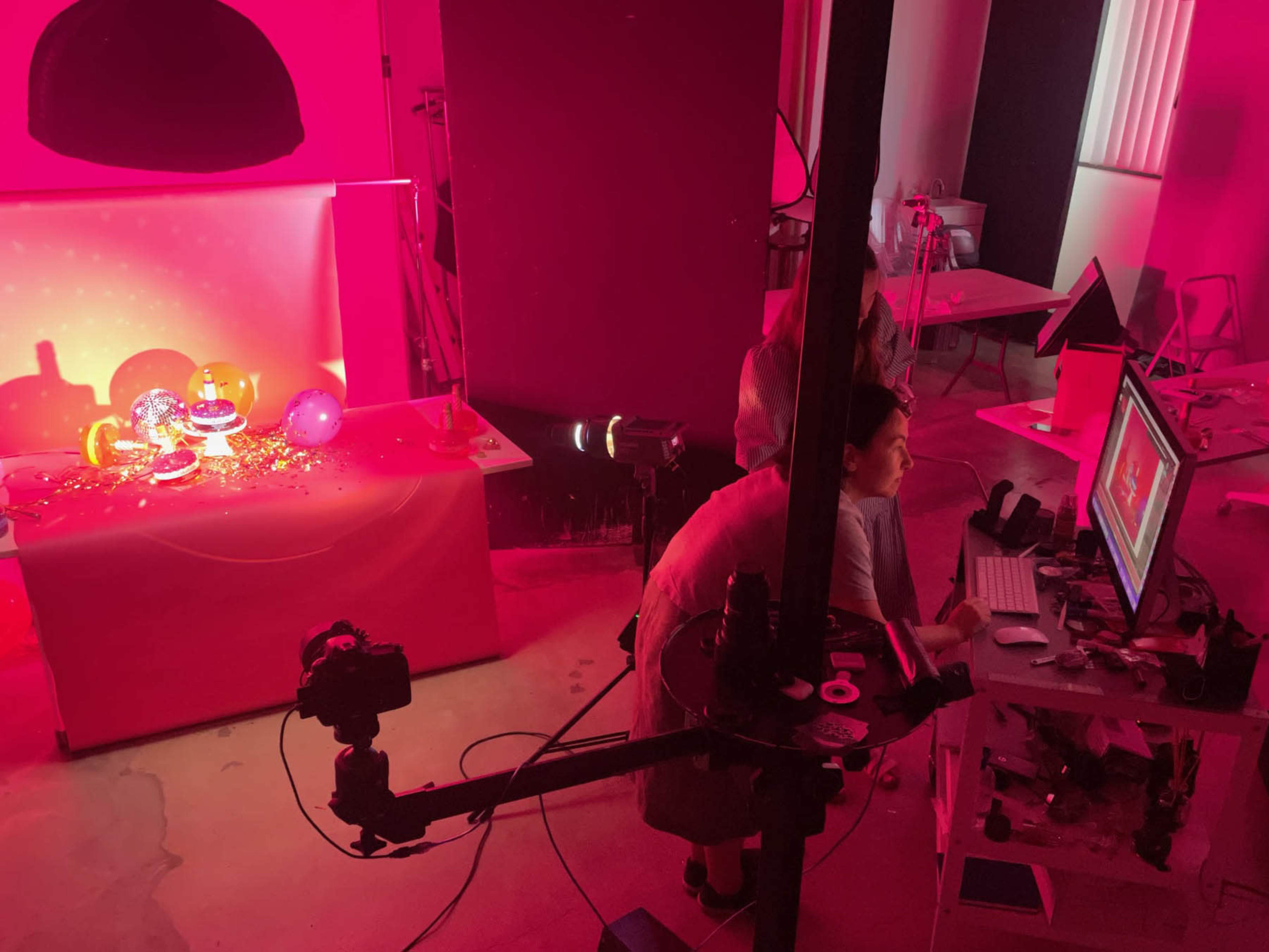 A brightly lit photography studio features a colorful setup with balloons and glassware, while a person sits at a computer monitoring the scene.