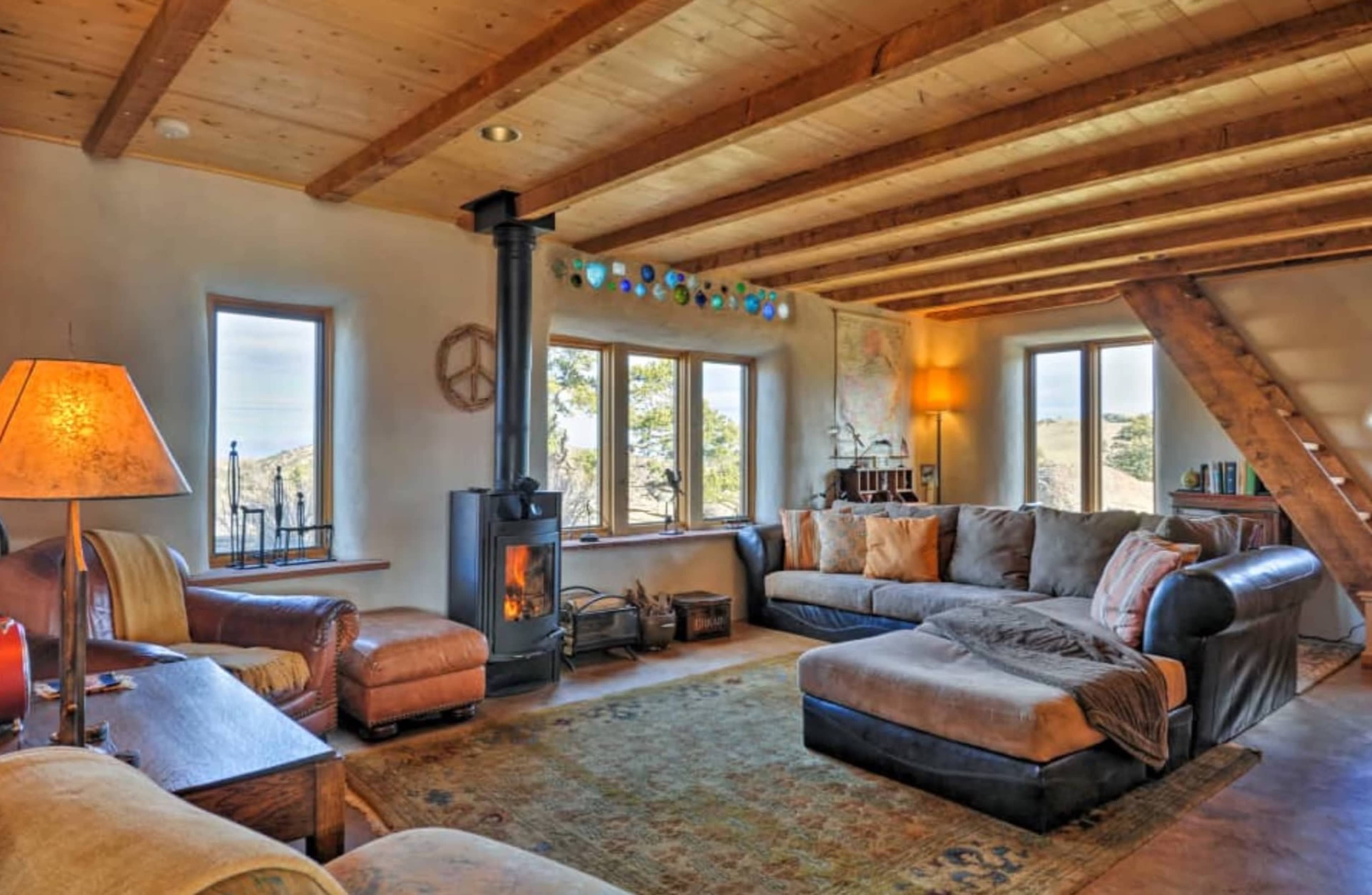 A cozy living room features a large sectional sofa, a wood stove, natural wood beams, and several windows providing views of the outdoors.
