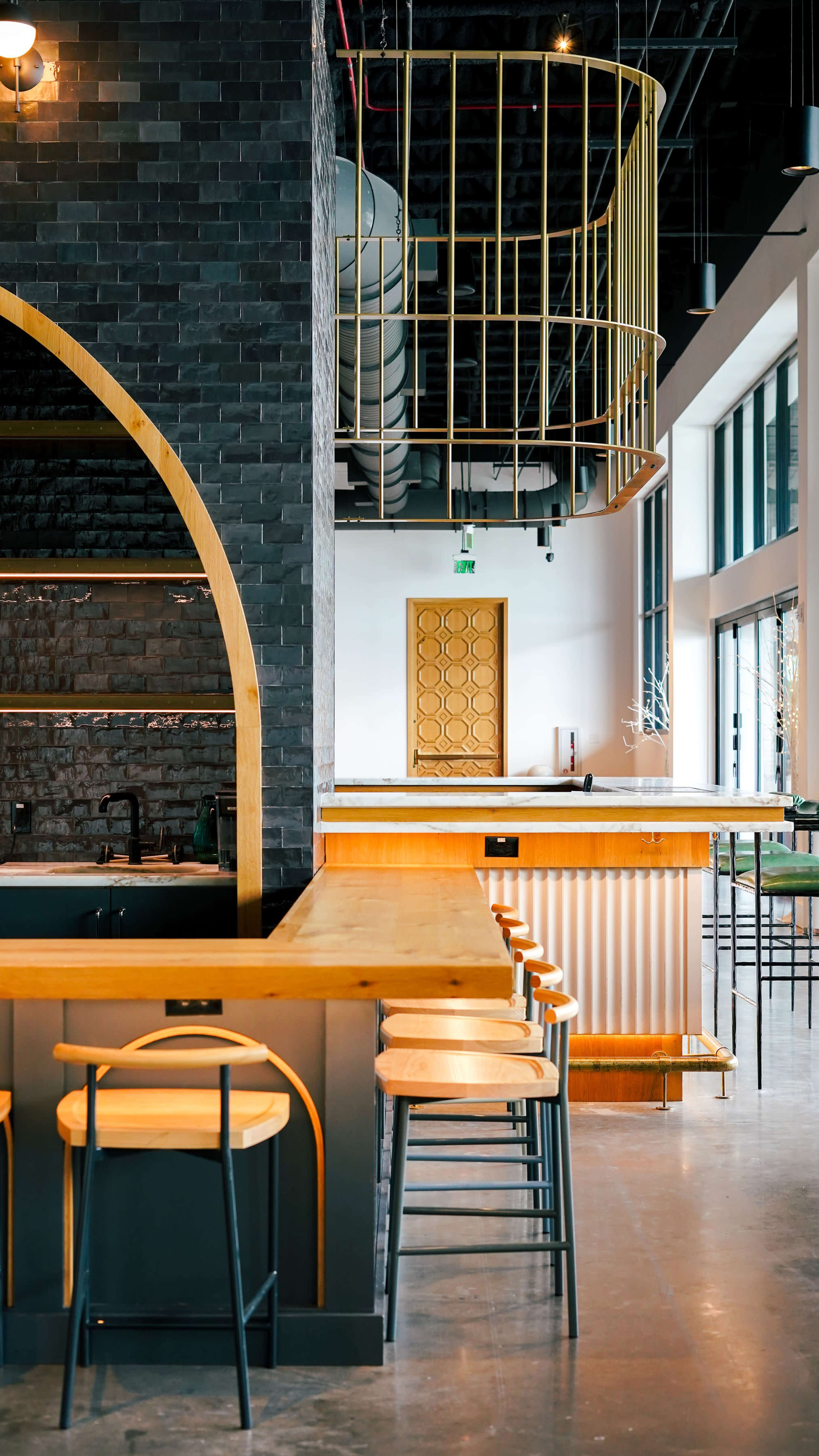 The image shows a modern interior of a cafe with a long wooden counter, black tiled walls, and a staircase featuring a gold railing in the background.