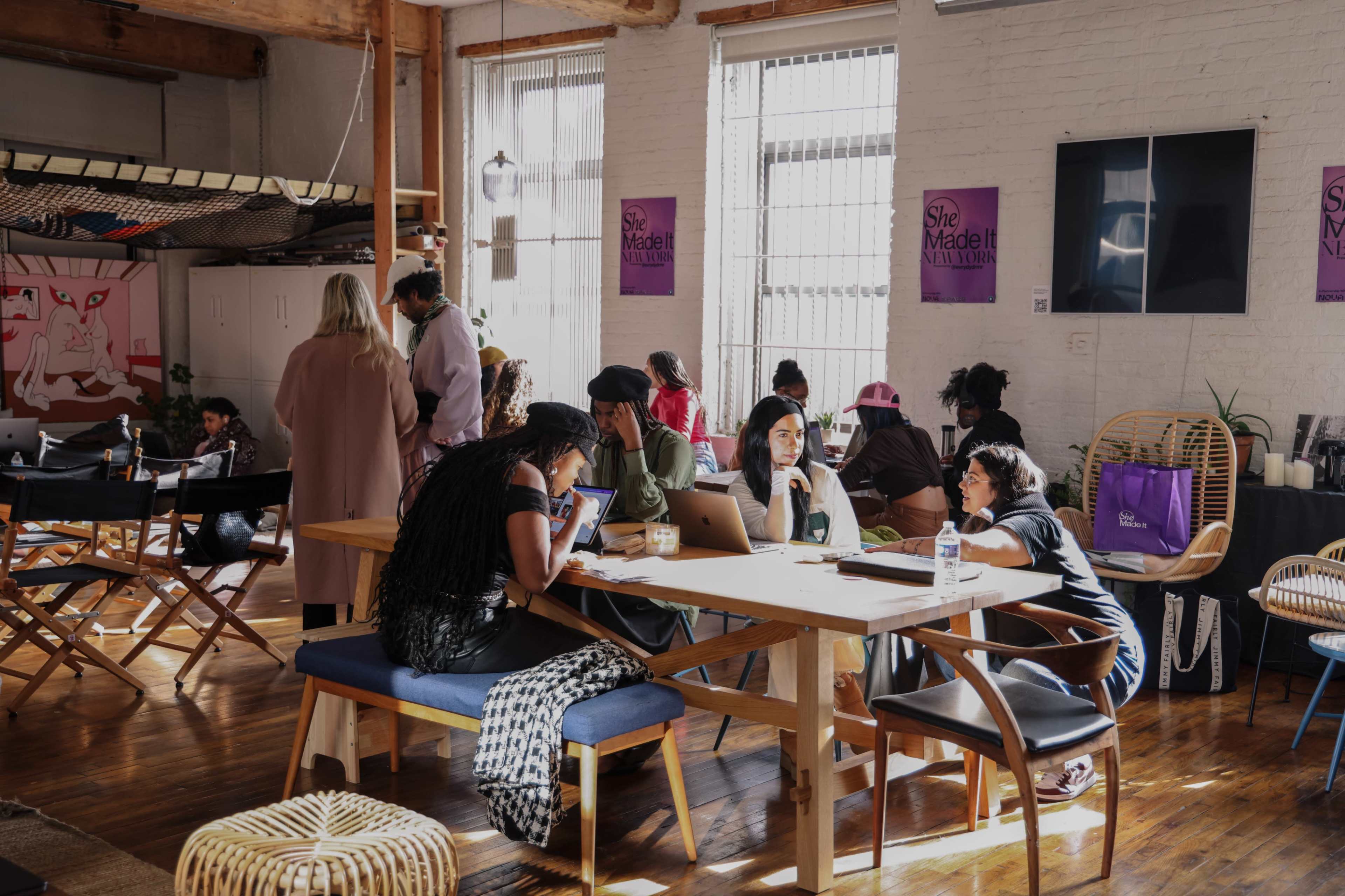 A group of people collaborates and engages in conversation around a large table in a brightly lit communal workspace.