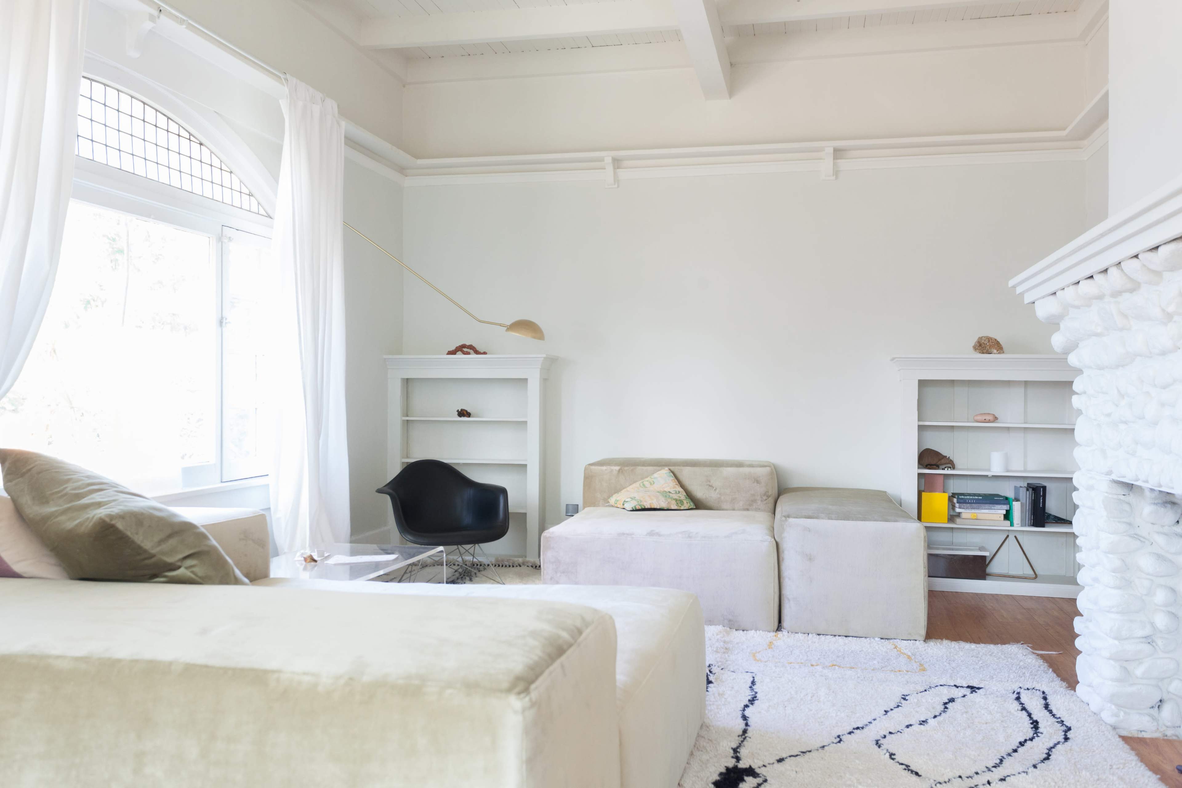 The image shows a modern, minimalistic living room with light walls, two sofas, a glass coffee table, and shelves displaying decorative items.