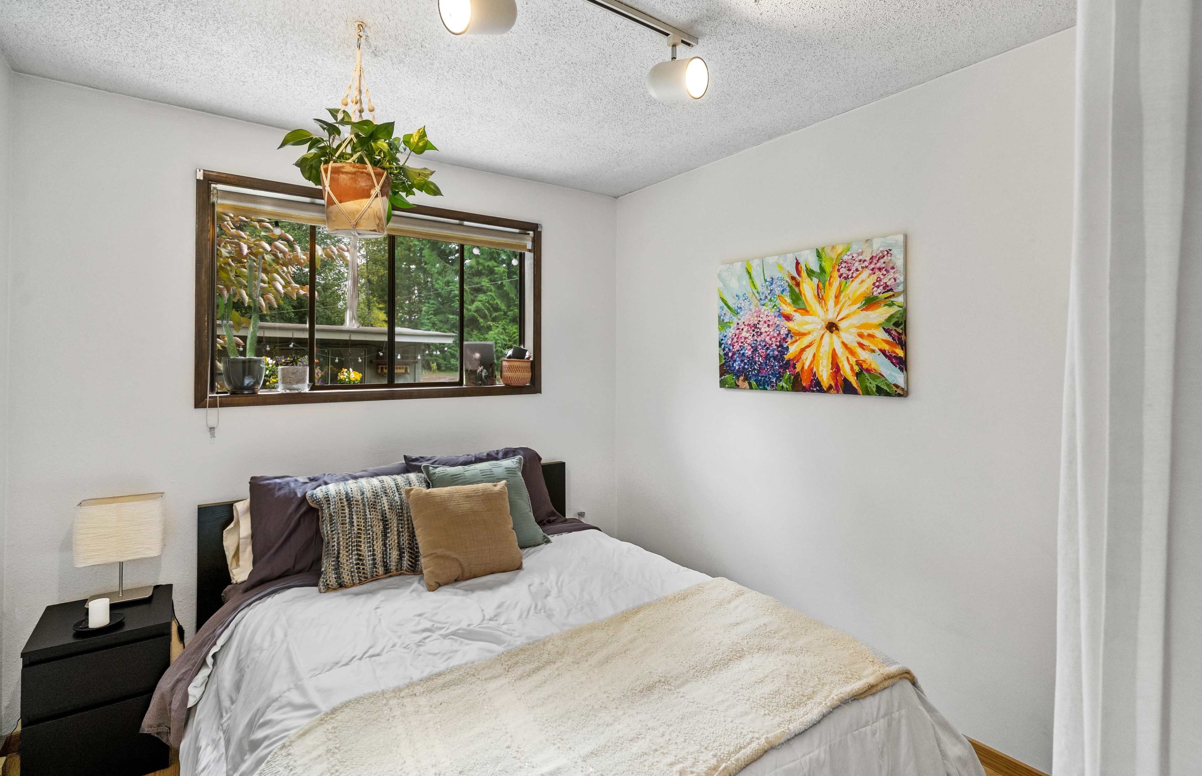 The image shows a well-lit bedroom featuring a bed with decorative pillows, a bedside lamp, and a window adorned with plants and art on the wall.