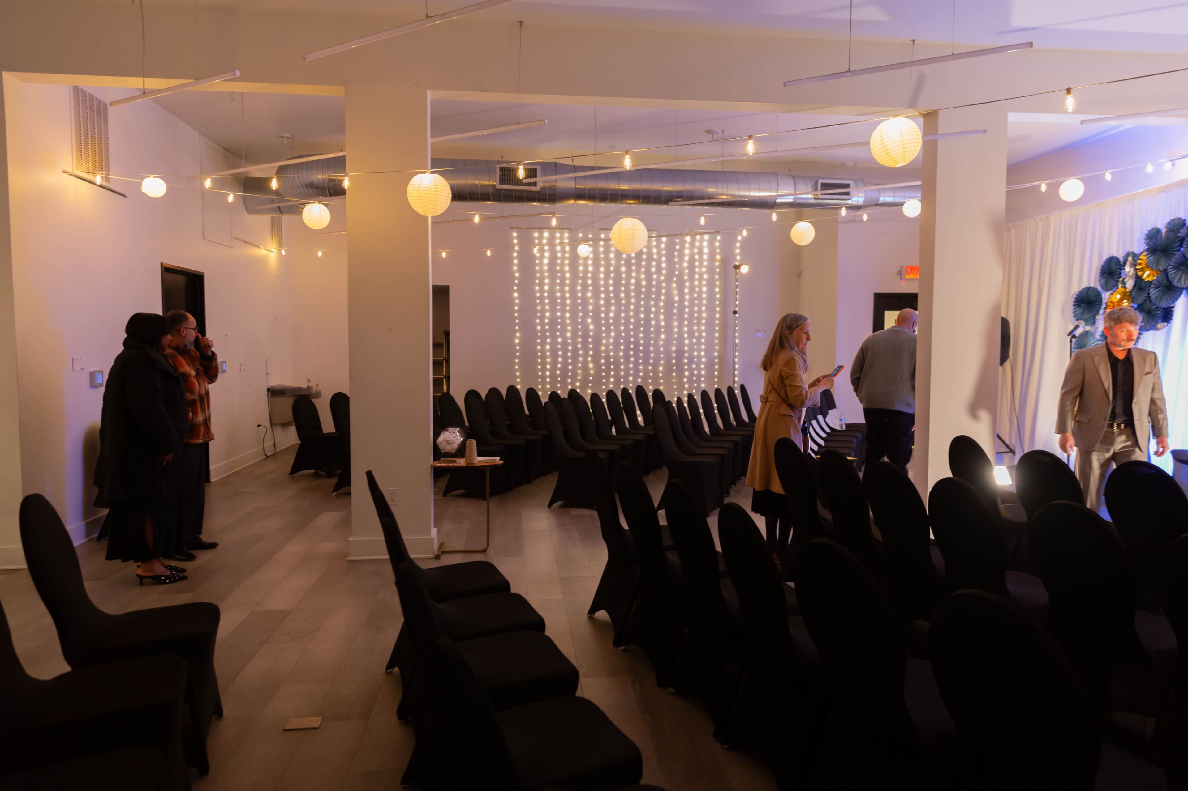 The image shows a spacious event area with rows of black chairs, softly lit with string lights and decorative lanterns, as a few people are moving around.