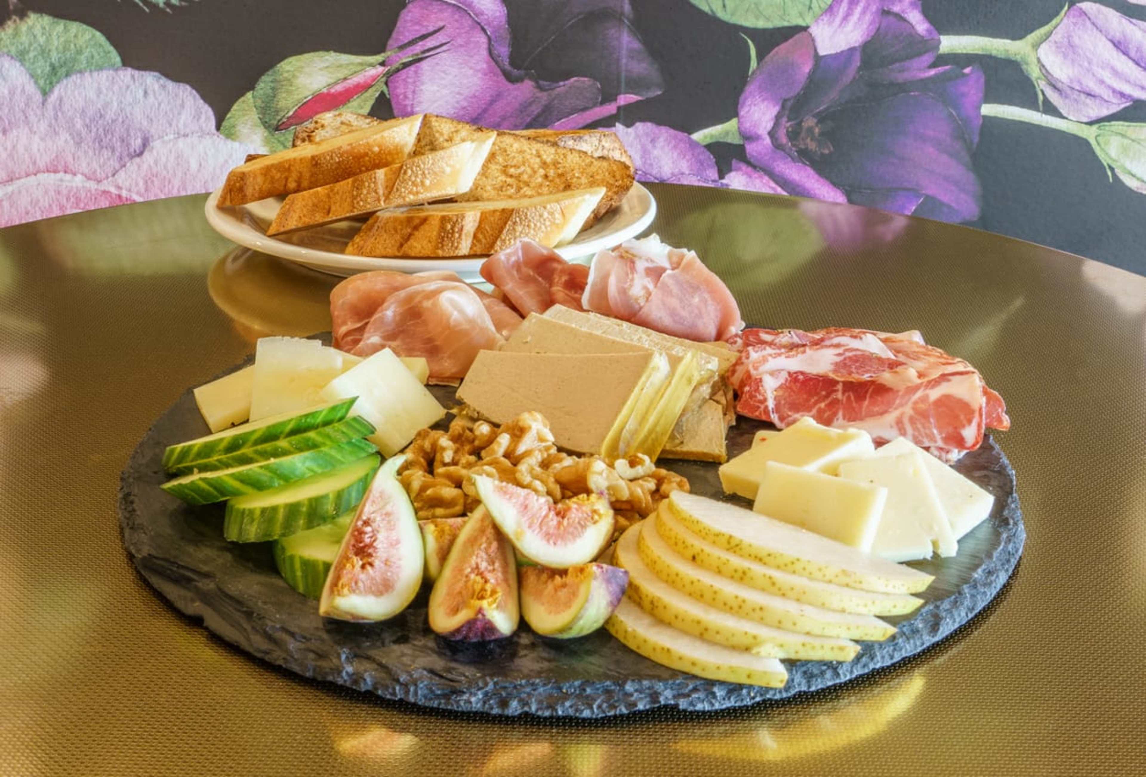 A platter of assorted cheeses, cured meats, fresh figs, and cucumbers is arranged on a slate board beside a plate of toasted bread slices.